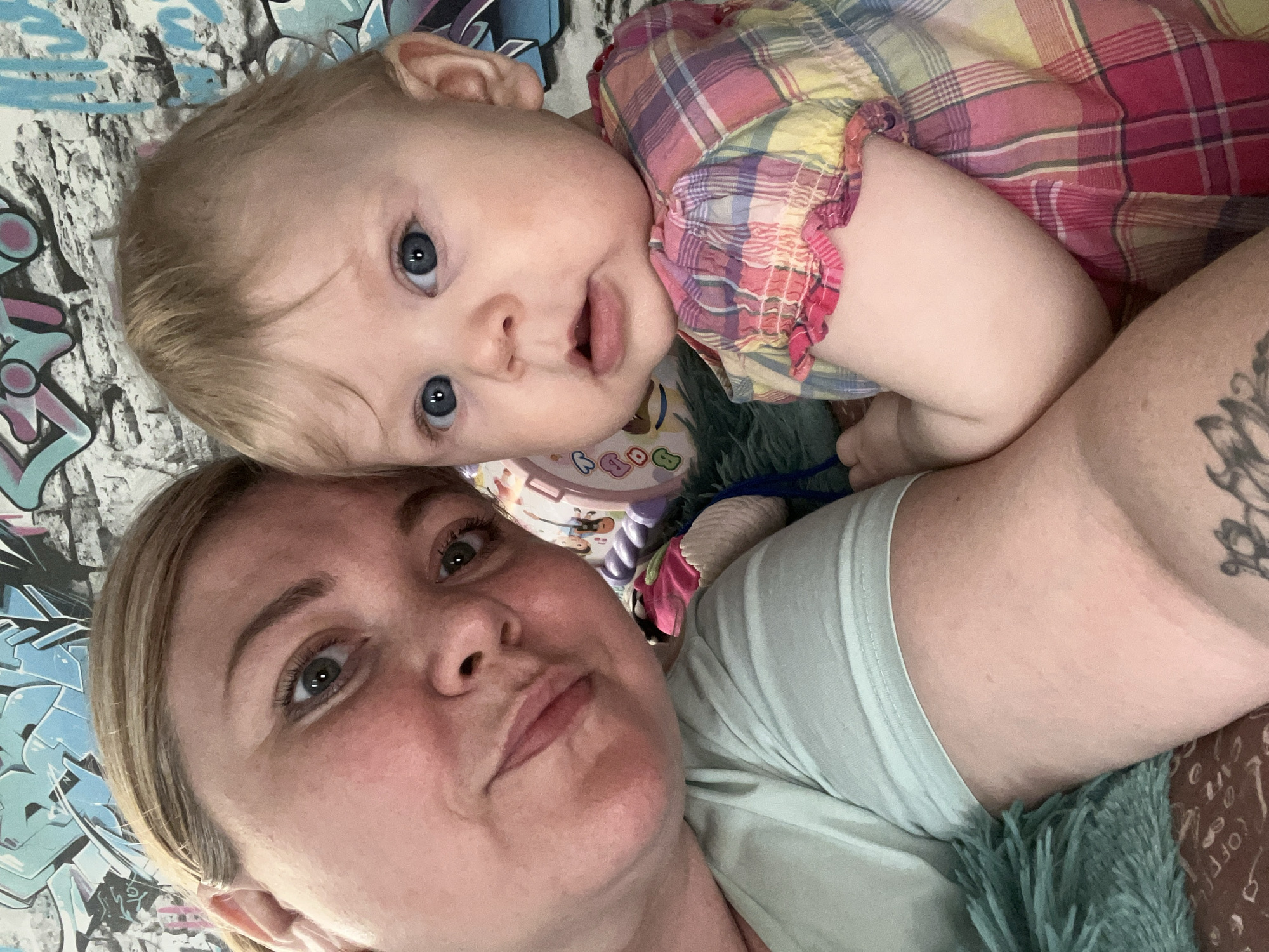 A woman and a baby lie close together. The baby is wearing a plaid dress and looks directly at the camera with an open mouth. The woman is wearing a light blue t-shirt. Part of a tattoo is visible on her arm. They are lying on a bed or surface with a blue and white graffiti-patterned sheet and a green fuzzy blanket.