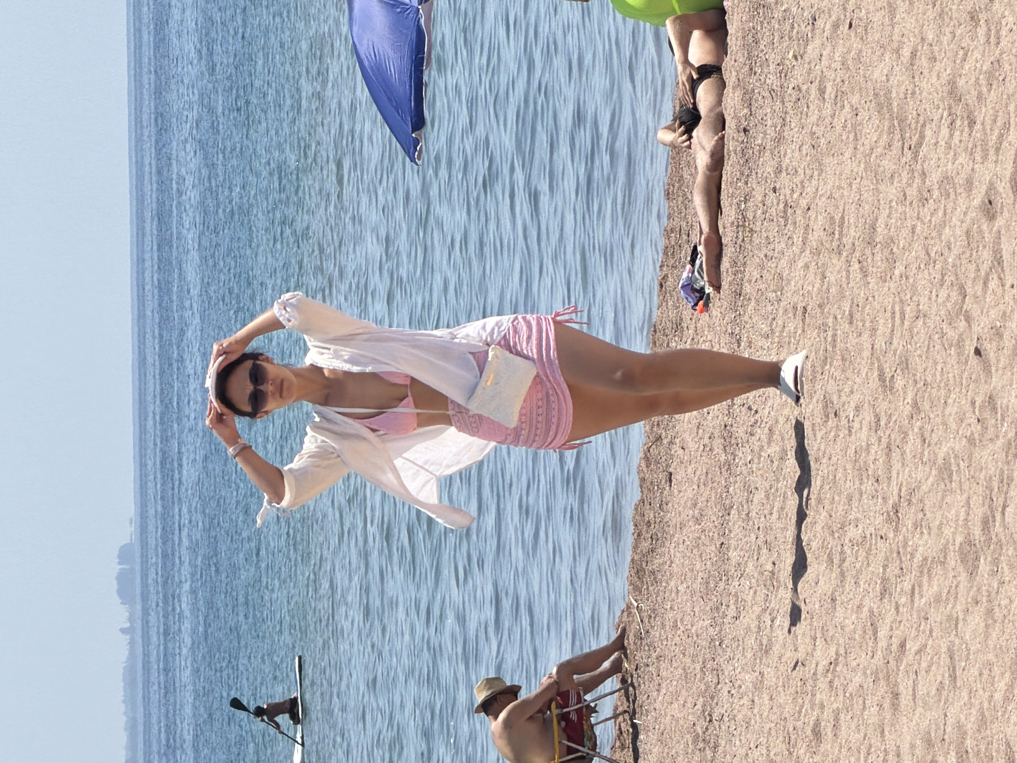 This is a full-shot, eye-level, outdoor photograph taken on a beach. In the foreground, a woman stands facing forward, wearing a pink bikini, a sheer white cover-up, and a small white purse across her body. She has her hands on her head, and she is wearing sunglasses and white shoes. To the left of the woman, a man sits in a folded chair, facing away from the camera, wearing a hat and red shorts. Further back in the water, a person is seen paddling a board. In the middle ground to the right, a blue umbrella is partially visible. In the background, the blue water stretches to the horizon under a clear sky. In the lower right, a person lies on the sand in black swimming trunks, with a green object behind them. The beach is composed of sand and small pebbles. The overall scene depicts a sunny day at a beach with several people engaged in various recreational activities.