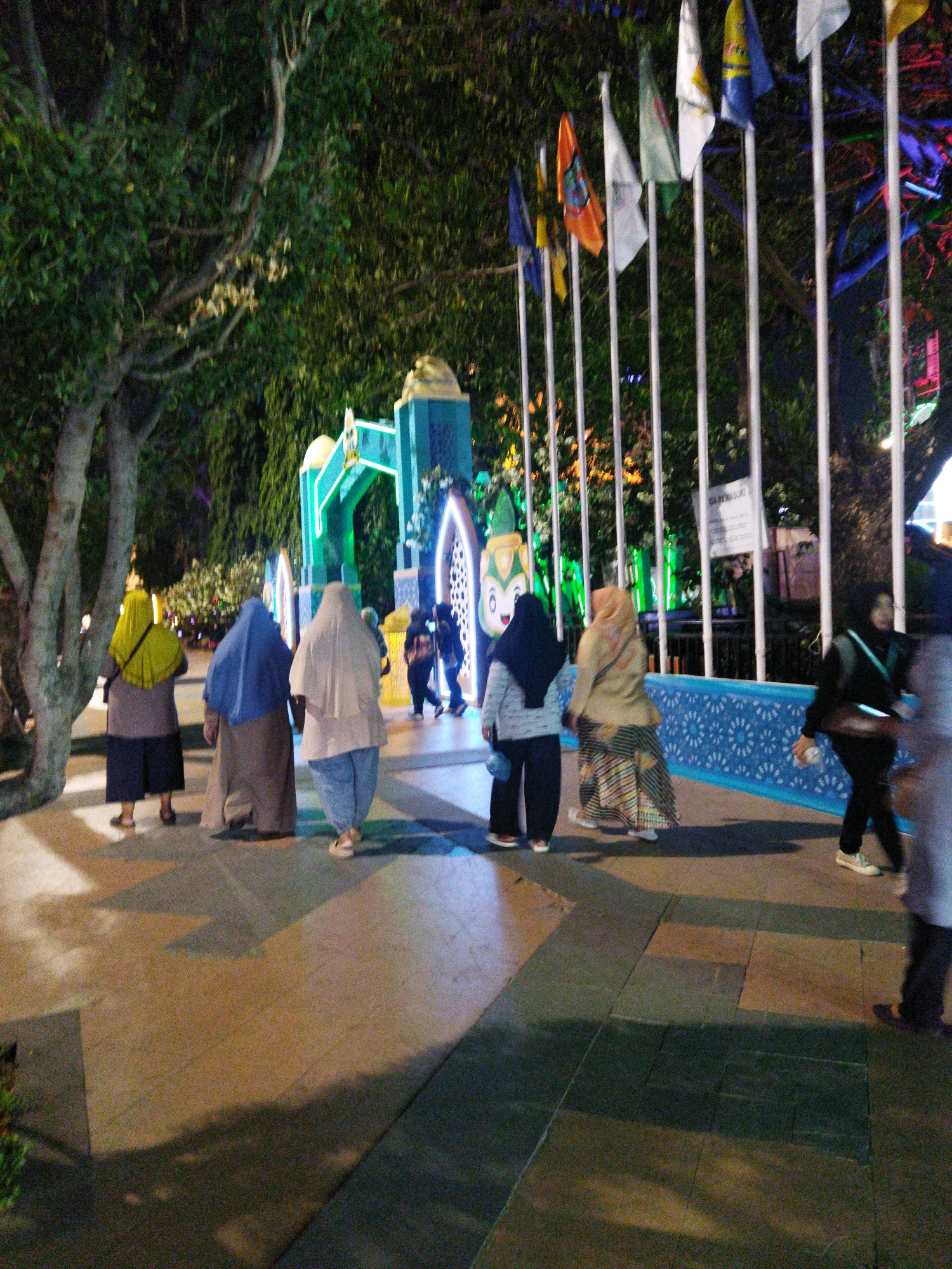 This is a nighttime, outdoor photograph taken in Semarang, Indonesia. The image depicts a group of people walking in a paved area in front of a decorative structure with an arched entrance. The structure appears to be illuminated with green lights and features decorative elements and flags on poles. Several individuals are seen walking away from the camera, most of them wearing headscarves and long garments, suggesting cultural attire. The lighting suggests it is evening or night, with artificial lights illuminating the scene and casting shadows on the ground. Trees are visible in the background. The ground is paved with large, rectangular tiles in varying shades of brown and gray, with geometric patterns formed by the arrangement of the tiles. In the distance, there are more lights and structures, hinting at a public space or event. The overall atmosphere is festive and public.