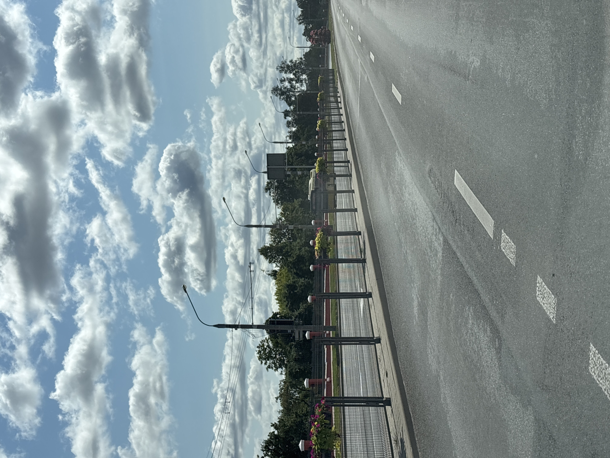 The image depicts a street scene in Minsk, Belarus, on a bright, cloudy day. The perspective is from the driver's seat of a vehicle looking forward along a multi-lane asphalt road. The road has visible white dashed lane markings. On the left side of the road, a paved sidewalk runs parallel to a metal fence. This fence is adorned with planters containing red and yellow flowers. Beyond the fence, trees and a red brick wall are visible. Streetlights with curved arms are spaced along the roadside, and overhead wires are also present. The sky is filled with scattered white and gray clouds. There are no people or other vehicles clearly visible in the immediate foreground or middle ground of the image. The overall impression is that of an urban thoroughfare on a pleasant day.