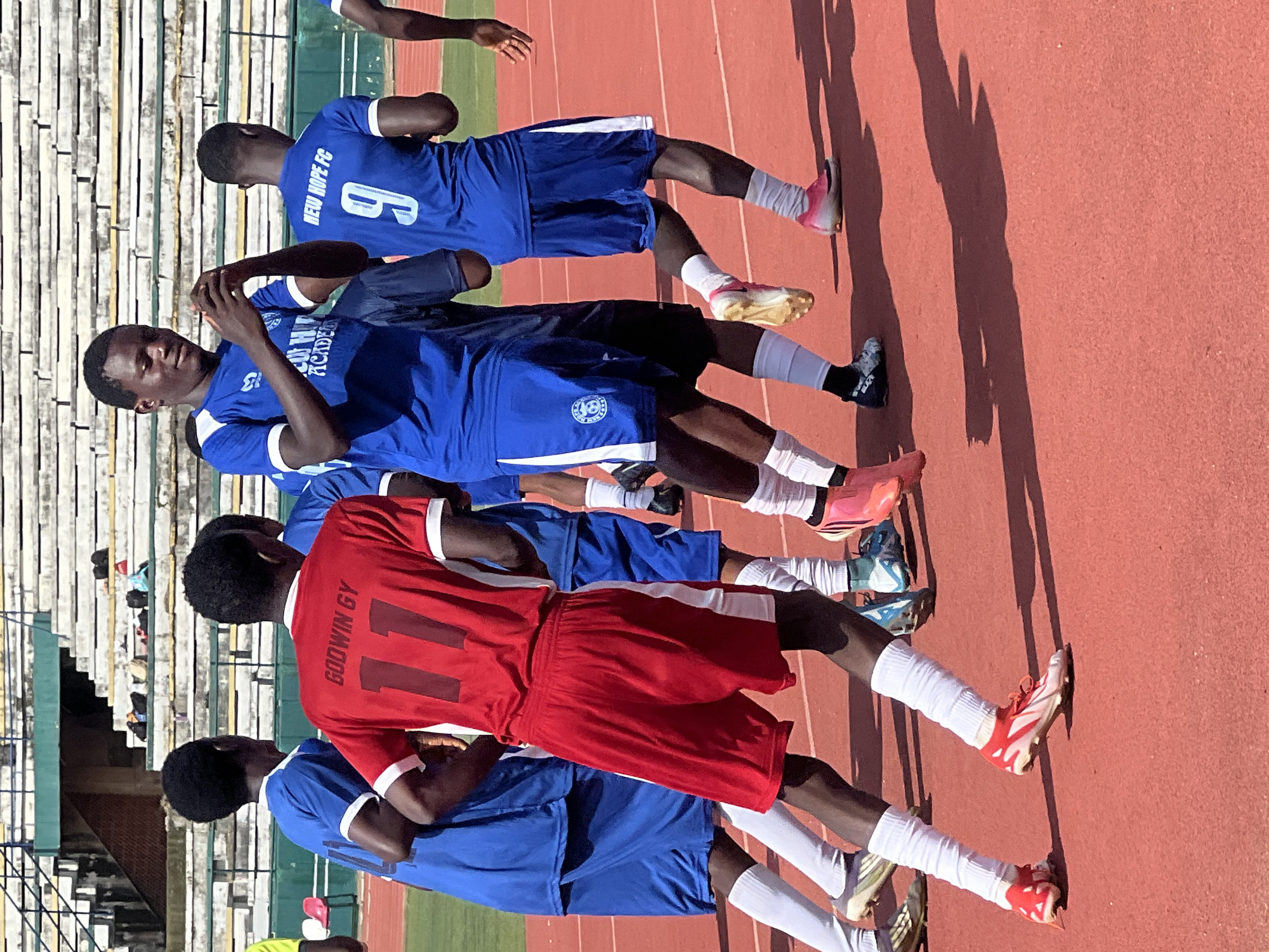 The image depicts a group of young male soccer players on a red synthetic track at the New Stadium in Nigeria. In the foreground, a player in a red jersey with the name "GODWIN GY" and the number "11" on the back is running, followed by other players in blue jerseys. The player in the red jersey is wearing white socks and red and white soccer cleats. Other players are also visible, some in blue jerseys with white trim, and numbers like "9" and "4" on their backs, and some with text that appears to read "NEW HOPE FC" and "ACADEMY". The environment includes stadium seating in the background, appearing somewhat weathered and with some bags visible on the steps. The ground is a bright red track, with shadows of the players cast on it, indicating a sunny day. The players are in motion, suggesting they are engaged in a game or training session.