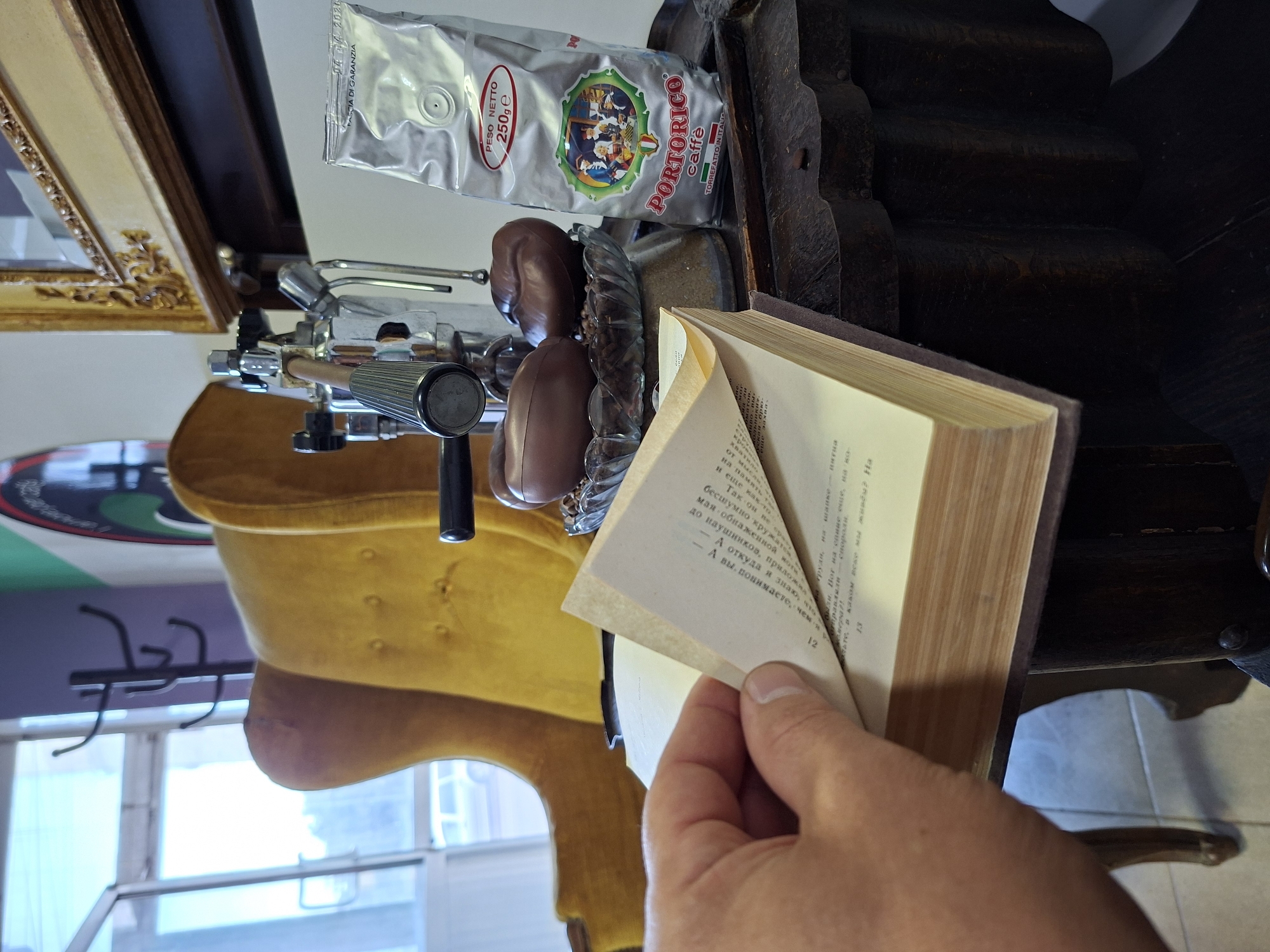 The image shows a close-up view of a book being held open by a left hand, with the pages turned. In the background, a vintage-style espresso machine is visible, along with a bag of "Portorico Caffè" coffee beans. There are also two chocolate confections shaped like coffee beans on a glass dish. The setting appears to be indoors, possibly a cafe or a home with a collection of coffee-related items and furniture. The overall impression is one of a cozy, European-inspired ambiance, possibly in a city like Chișinău, Moldova, known for its cafes.