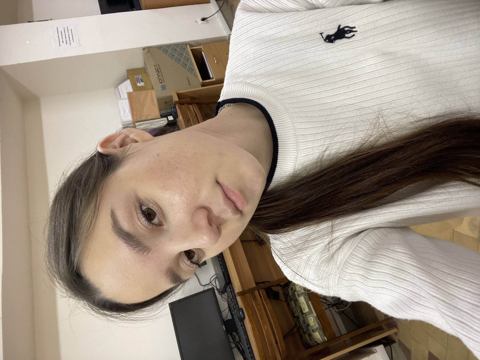 A young woman with light skin and brown hair is taking a selfie indoors. She is wearing a white, ribbed long-sleeved shirt with a small, black logo on the chest and a black trim around the collar. Her face is tilted towards the camera, revealing brown eyes and visible skin texture. The background includes a desk with a computer monitor, various cardboard boxes, and shelving. A paper with printed text is affixed to the wall in the upper left corner. The location is implied to be in Chaikovsky, Russia, based on the prompt.
