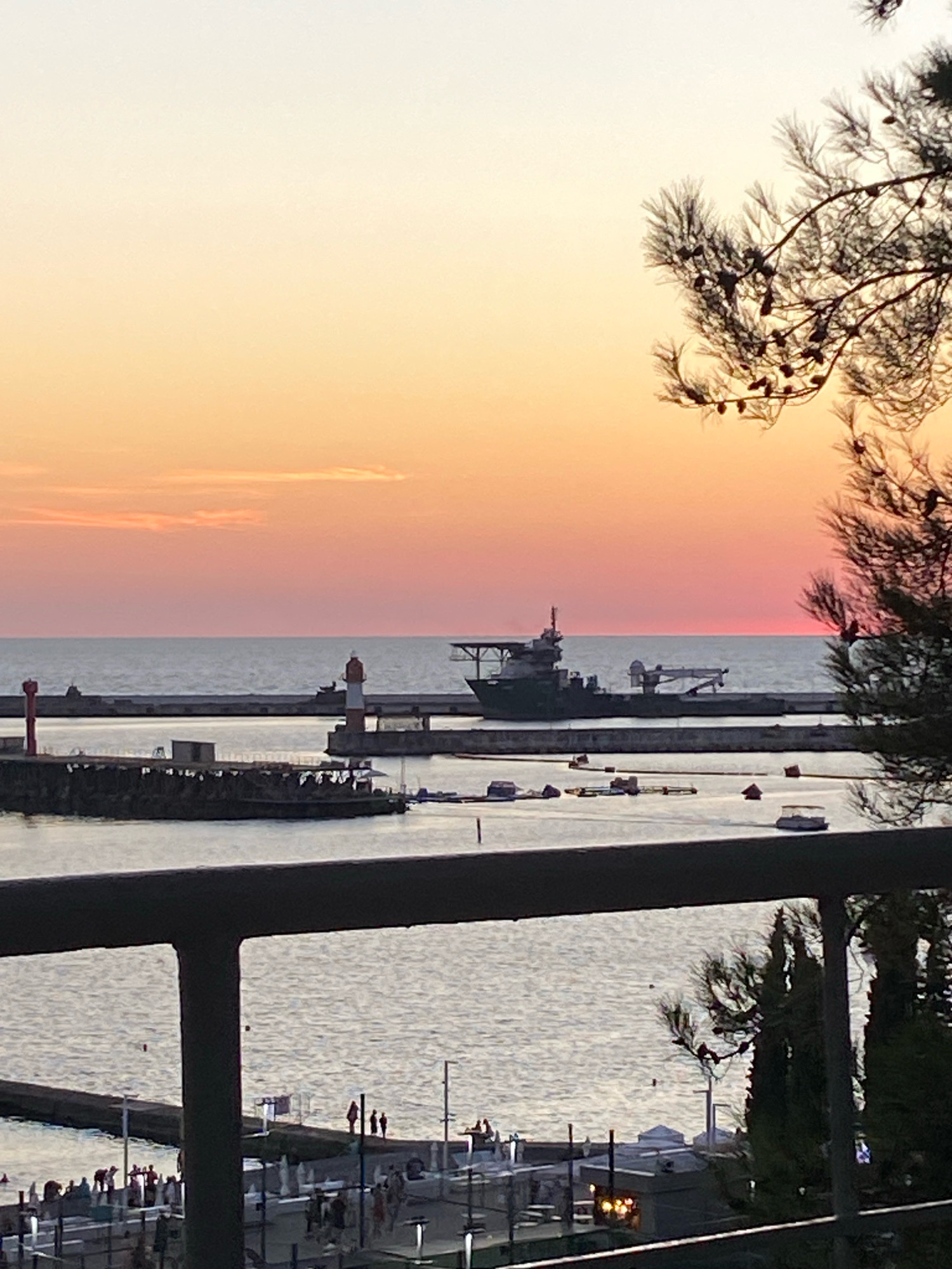 This image depicts a harbor scene in Sochi, Russia, during sunset. The sky displays a gradient of colors, transitioning from a pale yellow at the top to soft pinks and oranges near the horizon. A large, dark ship with a crane is visible in the middle ground, anchored in the calm water. To the left, a lighthouse with red and white stripes stands tall. The foreground is partially obscured by a dark railing and the silhouette of a pine tree branch with pinecones. In the lower part of the image, a promenade or boardwalk is populated with small figures of people, and several smaller boats are docked or moving in the water. The overall atmosphere is serene and coastal.