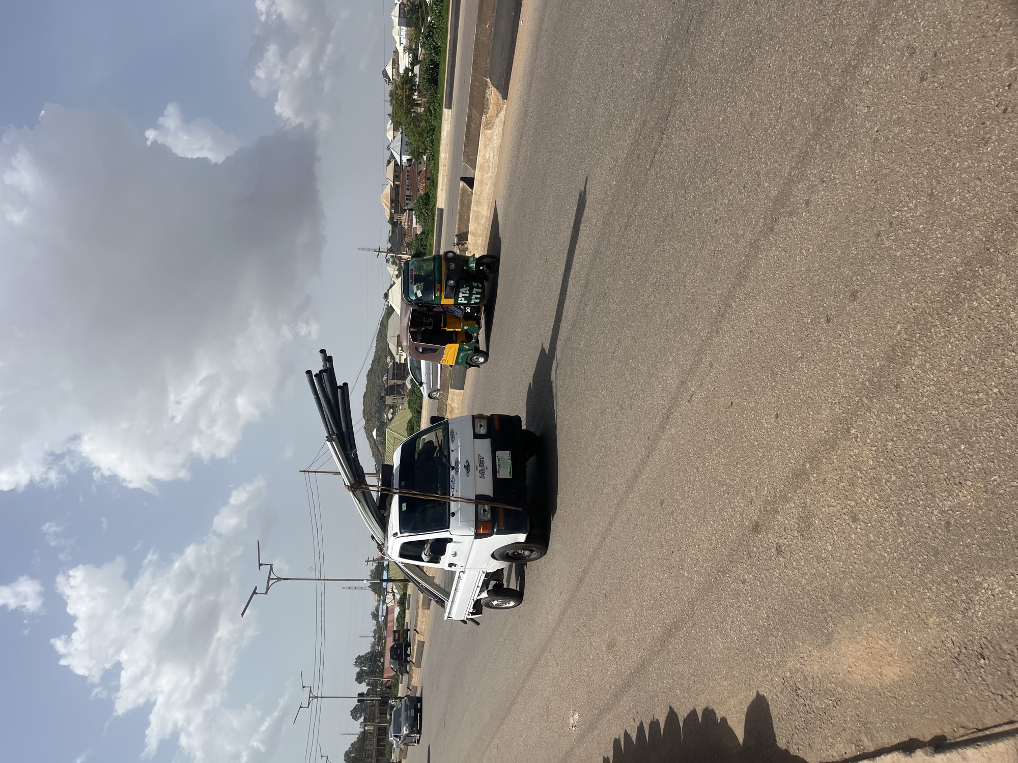 The image depicts a street scene in Gura Top, Nigeria, under a partly cloudy sky, suggesting it is daytime. The foreground is dominated by the side of a white pickup truck carrying a load of black pipes. The truck has "HIJET" written on its side, and a license plate. Behind the truck, a green and yellow auto-rickshaw, also known as a "Keke Napep," is visible. The auto-rickshaw has text on its side, including "PTA 4775". The background reveals a residential area with houses and buildings, some with corrugated iron roofs, set against a backdrop of green vegetation and a hill. Streetlights are visible along the road. The overall scene suggests a normal day of transportation and activity in a Nigerian town.