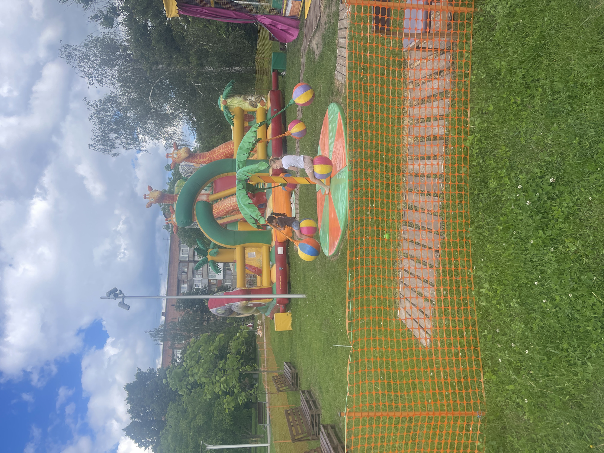 An outdoor playground features a large, inflatable jungle-themed structure with slides and climbing areas, adorned with giraffe and monkey motifs. In the foreground, two children are seated on a circular, rotating platform with colorful balls hanging from a palm tree structure. One child, wearing a white t-shirt and light-colored shorts, faces away from the camera, while the other, in a dark t-shirt and jeans, is partially visible. The playground is surrounded by a green grassy area, with wooden benches placed to the left. An orange safety net fence is in the immediate foreground, with a wooden walkway visible behind it. In the background, trees and a multi-story brick building are visible under a partly cloudy sky. The scene appears to be in a city park or recreational area.