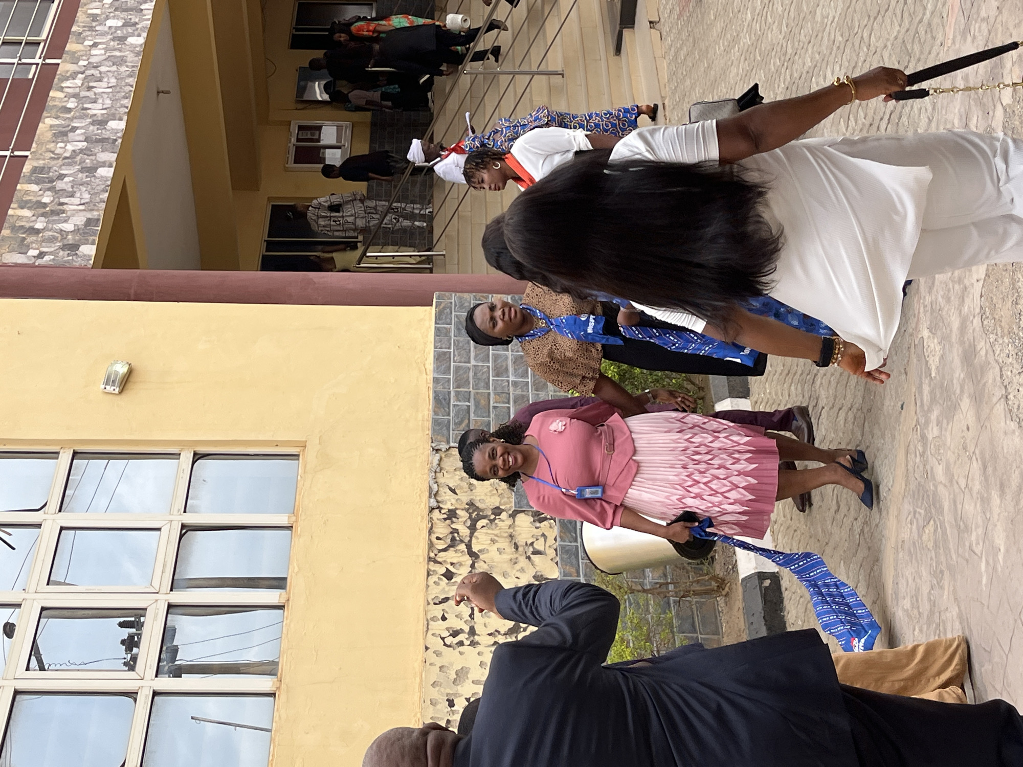 A group of people stand outside a building in Abuja, Nigeria. One woman wears a pink dress and smiles, while another wears white clothing and holds a walking stick. A man in a dark suit points towards the group. Several other people are visible in the background, some on steps and others near the building's facade. The building has windows, a yellow exterior, and a textured section. The ground is paved.