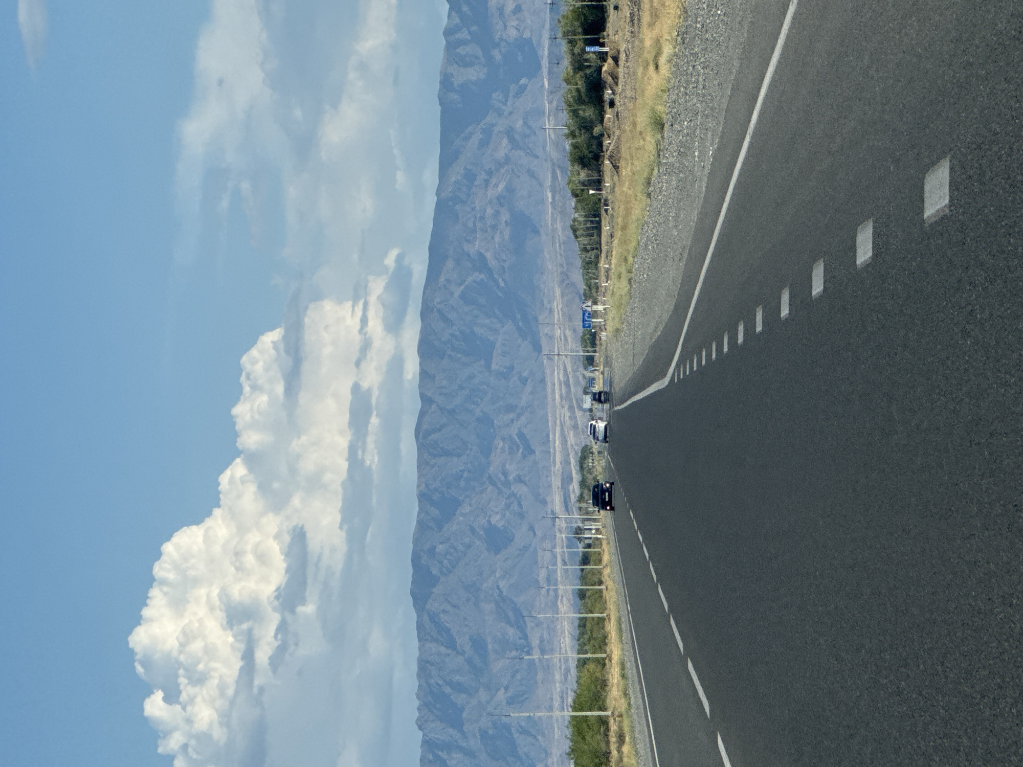 The image depicts a highway stretching towards a distant mountain range under a partly cloudy sky. The road is asphalt with white lane markings. Several cars are visible on the road, moving away from the viewer. The landscape on either side of the road consists of dry, grassy vegetation and scattered trees. A line of utility poles runs along the right side of the road. In the background, a large, rugged mountain range dominates the horizon, its slopes etched with shadows and texture. The sky is a vibrant blue with large, puffy white clouds. The scene appears to be a view from a vehicle traveling on a highway in a rural or semi-arid region. Given the reference to Üşaral, Kazakhstan, this is likely a highway leading into or out of that area.