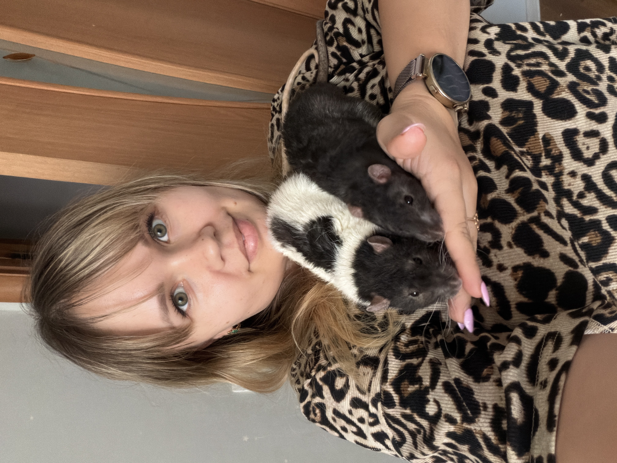 A person is holding two pet rats. The person has blonde hair, green eyes, and is wearing a leopard print shirt. They also have a watch on their left wrist and manicured nails. One rat is black, and the other is black and white. The background includes a wooden object. The location is unspecified.