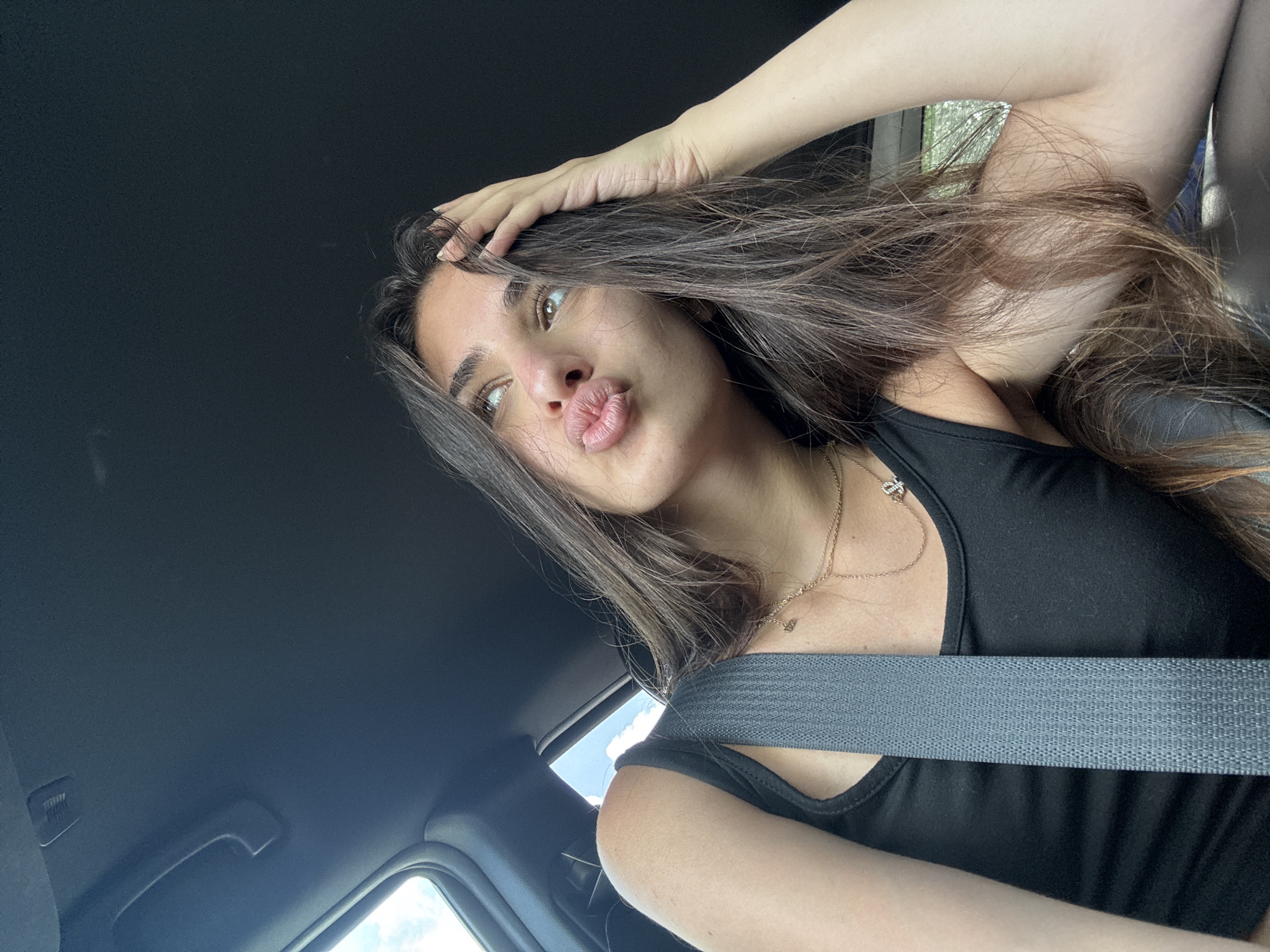 A young woman is inside a car, wearing a black tank top and a seatbelt. She is puckering her lips and has one hand on her head, which is covered with long brown hair. The car's interior, including the roof and a window, is visible. The location is likely Delray Beach, United States of America.