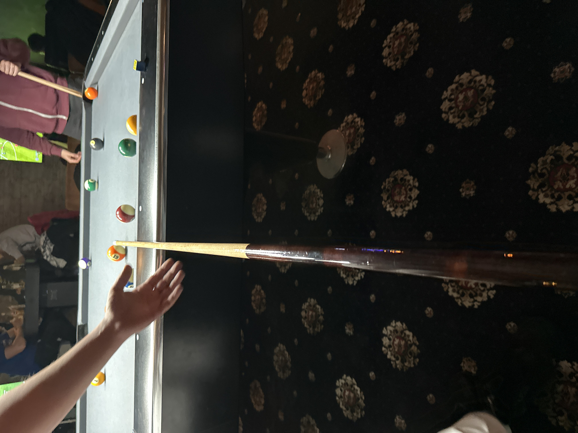 The image depicts a person aiming a pool cue at a pool table. The pool table has multiple billiard balls on it. The background is a wall with a patterned design and a pool cue holder. There is no indication that the image was taken in Plovdiv, Bulgaria.