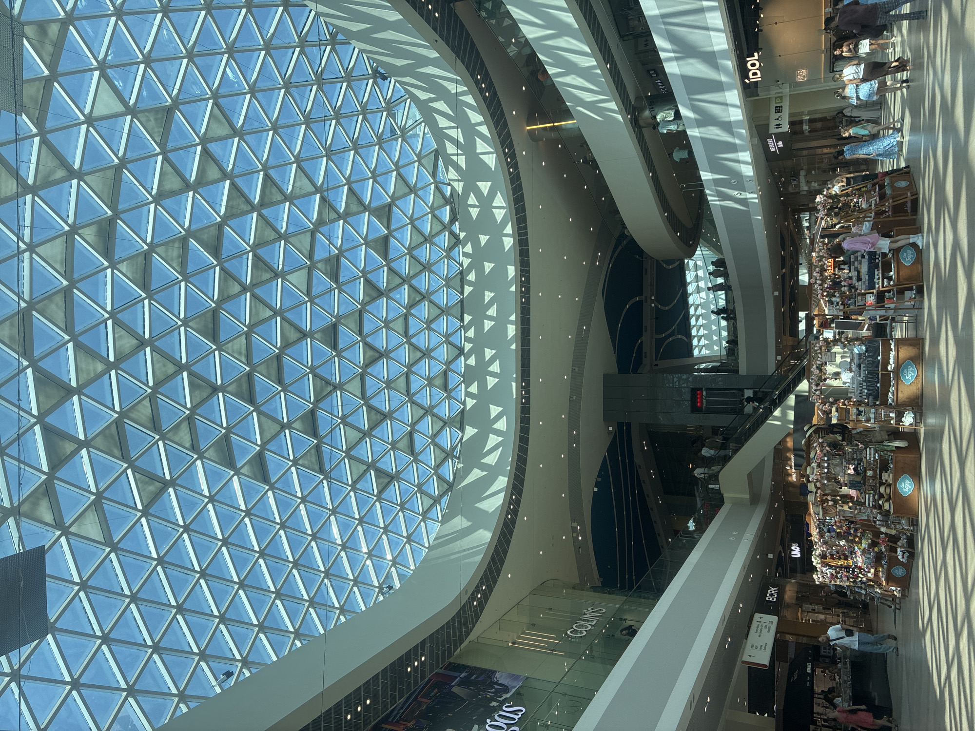 The image captures the interior of the "More Mall" in Sochi, Russia, a large shopping center characterized by its modern architecture. The most prominent feature is the expansive, geodesic dome-like glass roof that bathes the space in natural light, creating intricate patterns of light and shadow on the floors and interior structures.
The mall features multiple levels with curved balconies and walkways, interconnected by escalators and staircases. Various shops are visible along the different levels, with some signage readable, such as "COLINS," "BORK," "UNO," and "IDOL."
On the ground floor, several retail displays are set up, including a stall with a variety of items like hats, bags, and other accessories, suggesting a marketplace or temporary vendor area. People are seen browsing and walking through the mall, engaged in typical shopping activities. A few individuals are visible on the escalators and upper levels. The environment appears clean and well-lit, with modern lighting fixtures supplementing the natural light from the dome. The overall impression is that of a bustling, contemporary retail environment within a geometrically striking architectural design.