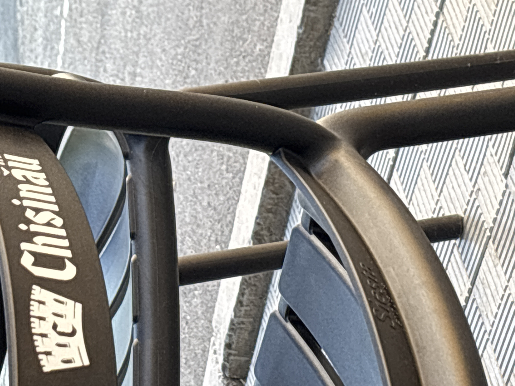 The image shows a close-up of a dark-colored chair, likely made of plastic or metal, with a logo on its backrest. The logo features a stylized castle with the text "Chisinau" written in white capital letters next to it. The chair is positioned outdoors, with a paved ground surface and a curb visible in the background. The lighting suggests it is daytime. The chair appears to be part of an outdoor seating arrangement, possibly at a cafe or public space.