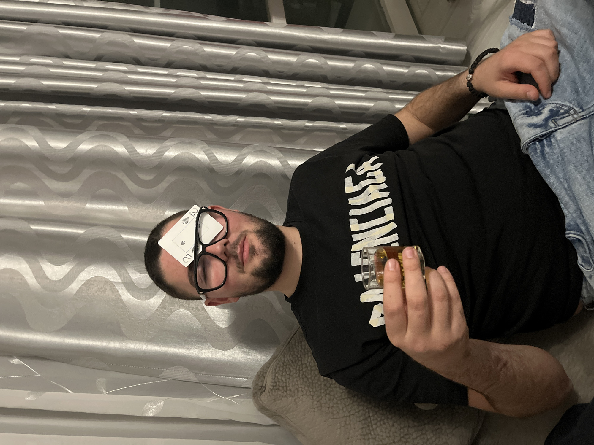 A man is lying down, possibly on a couch or bed, indoors. He is wearing black-rimmed glasses, a black t-shirt with white lettering, and a bracelet on his right wrist. He has a playing card stuck to his forehead. He is holding a shot glass filled with amber liquid in his right hand. A denim jacket is partially visible next to him. The background includes a silver curtain with a wavy pattern. It is unknown if the location is Burgas, Bulgaria.