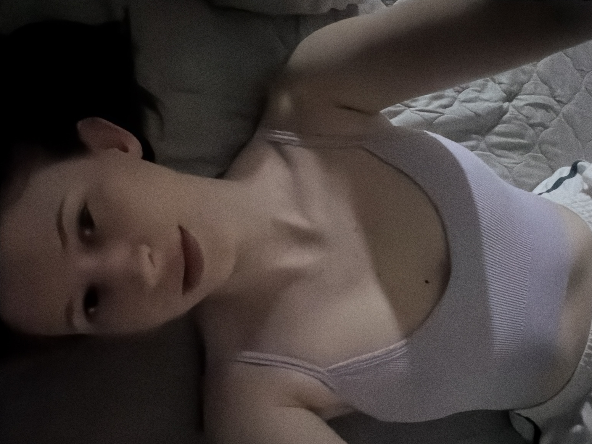 A person is lying on their back on a bed, with their arms raised above their head. The person is wearing a light purple tank top and white shorts. The bed is covered with a gray quilted blanket. The lighting in the room is dim, and the image has a grainy texture. The image is a selfie taken from a high angle, looking down at the person. There is no discernible city or location information in the image.