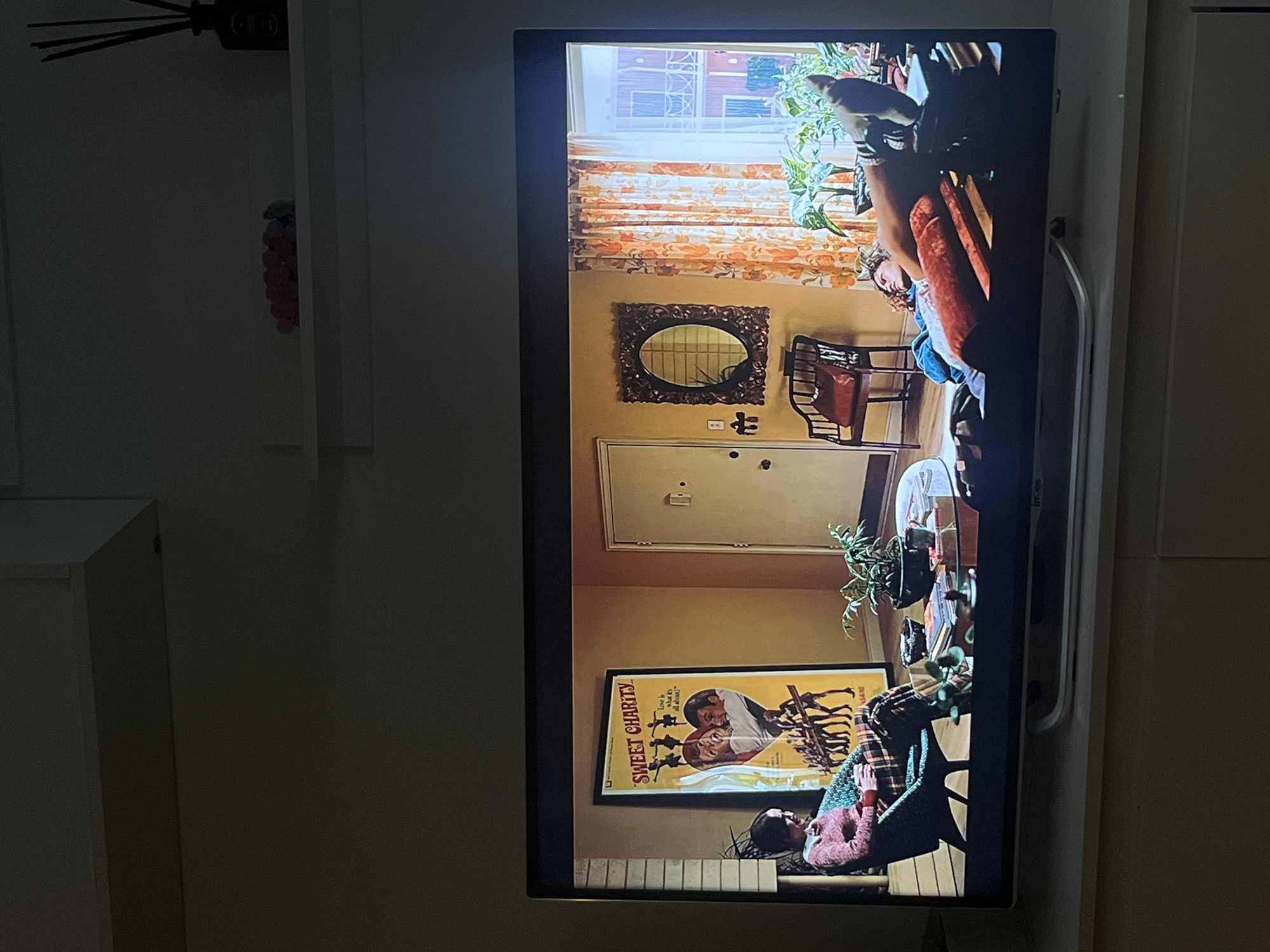 A television displays a scene from a film, featuring a room interior. The interior has patterned curtains, a mirror, a door, a poster that reads "Sweet Charity," furniture including chairs, and potted plants. A person is visible in the scene, resting on a surface near the poster. The scene appears to be from the film "Sweet Charity." It is impossible to determine from this picture if it is located in Madrid, Spain.