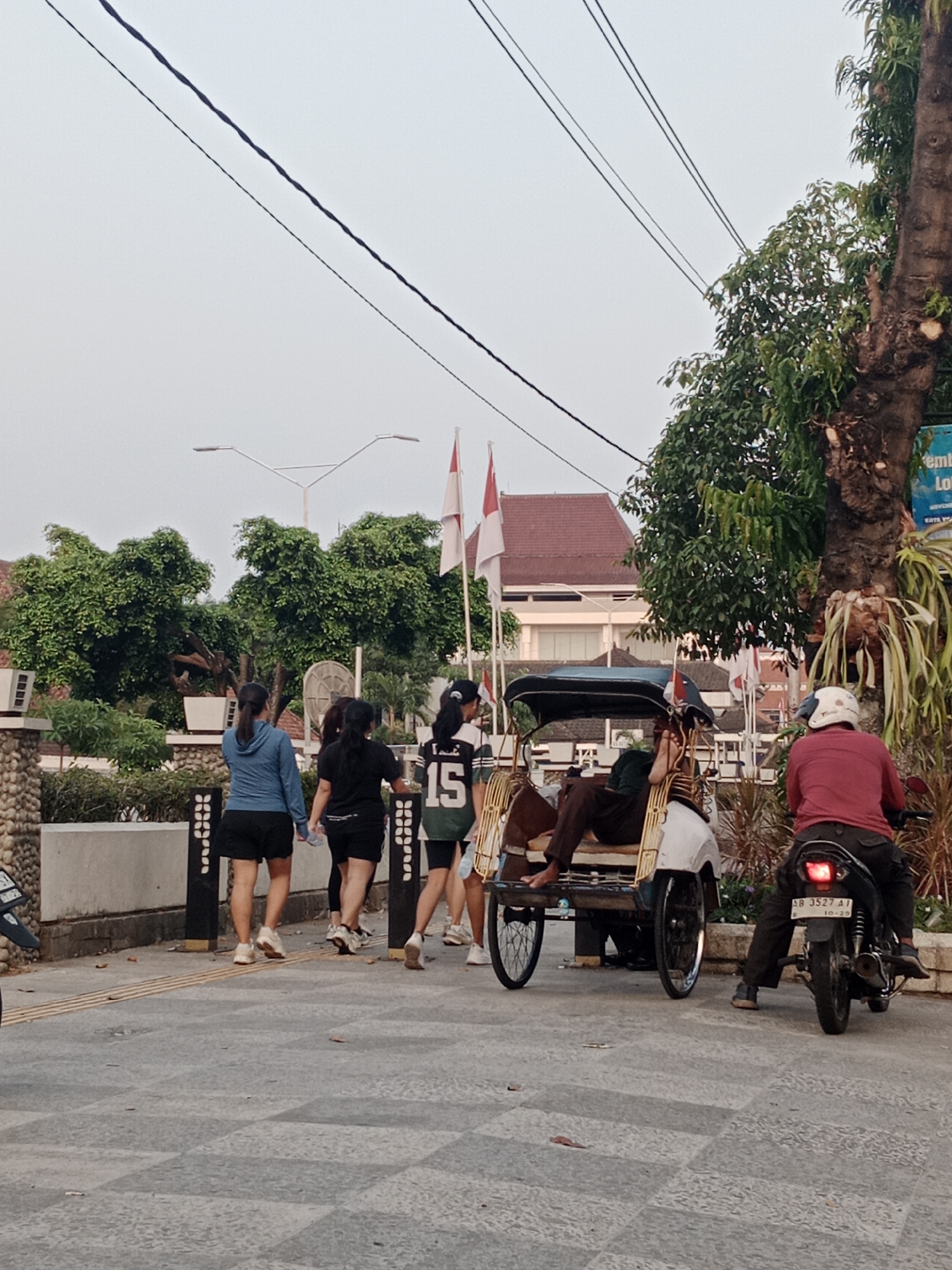 In Yogyakarta, Indonesia, an outdoor scene depicts a street in the late afternoon. A becak, a three-wheeled passenger vehicle, is parked on the right side of the frame, with a person reclining inside and another person sitting on the front. A motorcyclist wearing a red shirt and a white helmet is stopped behind the becak, facing away from the camera. In the background, several people are walking, and Indonesian flags are visible. The area appears to be a public space with trees, buildings, and a paved walkway.