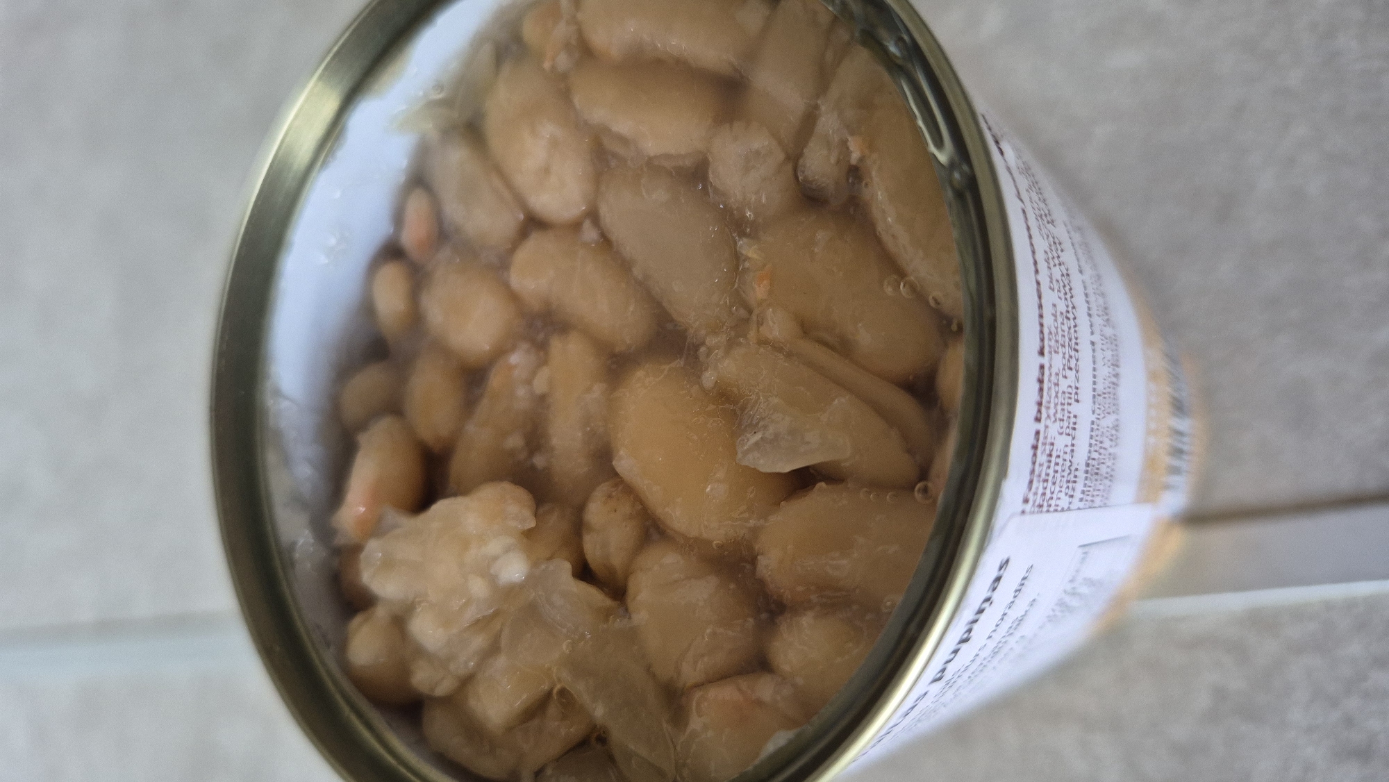 The image shows a close-up view of an open can of white beans. The beans are submerged in liquid and appear plump and slightly translucent. Some of the beans show a creamy white color, while others have a more yellowish hue. Bubbles are visible within the liquid, suggesting the beans are preserved. The can's rim is visible at the top of the image, and a portion of a label with text is visible on the side of the can. The background appears to be a neutral, light-colored surface. There are no people or specific environmental elements of Krauja, Latvia identifiable in this image.