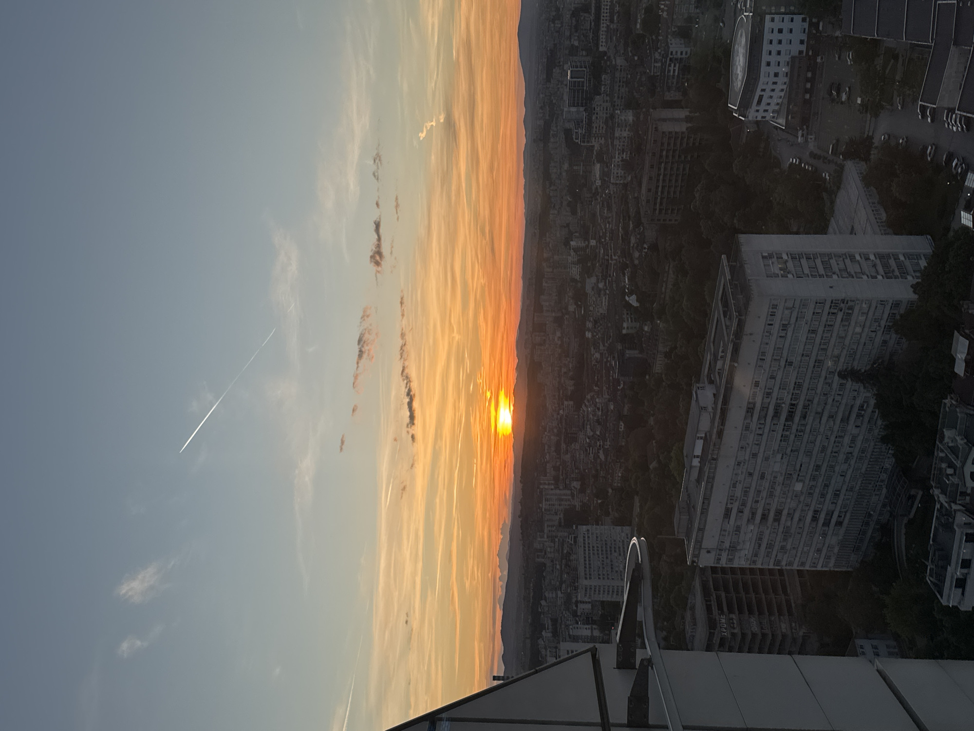 The image shows a sunset over the city of Sofia, Bulgaria, viewed from a high vantage point, possibly a rooftop. The sky transitions from blue to orange and yellow near the horizon, with streaks of clouds. The sun is positioned on the horizon line. Below the horizon, a cityscape of Sofia extends into the distance, appearing dark in contrast to the bright sky. Numerous buildings are visible, varying in height and style. At the bottom of the image, there are rooftop elements like railings. A contrail from an aircraft is visible in the blue part of the sky.