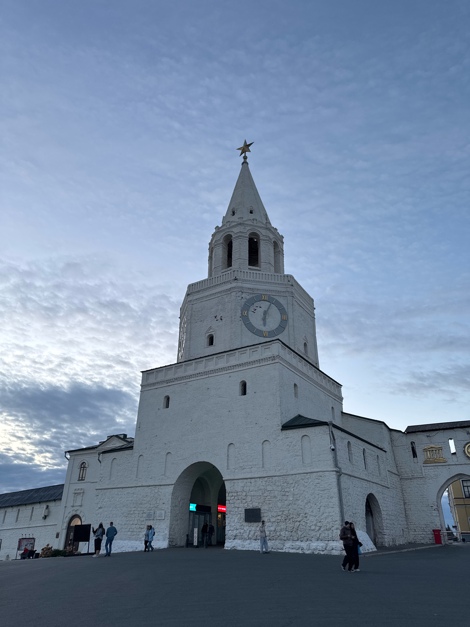 The image depicts the Söyembikä Tower, a prominent landmark in the Kazan Kremlin, Kazan, Russia. The tower is a white, multi-tiered structure with a clock face and a golden star atop its spire. The sky above is overcast with a mix of white and gray clouds. In the foreground, several people are walking on a paved area in front of the tower. Some are standing in small groups, while others are walking individually or in pairs. The base of the tower features large arched entrances, some of which appear to lead to shops or establishments. The overall scene suggests a public space with moderate foot traffic under a muted sky.