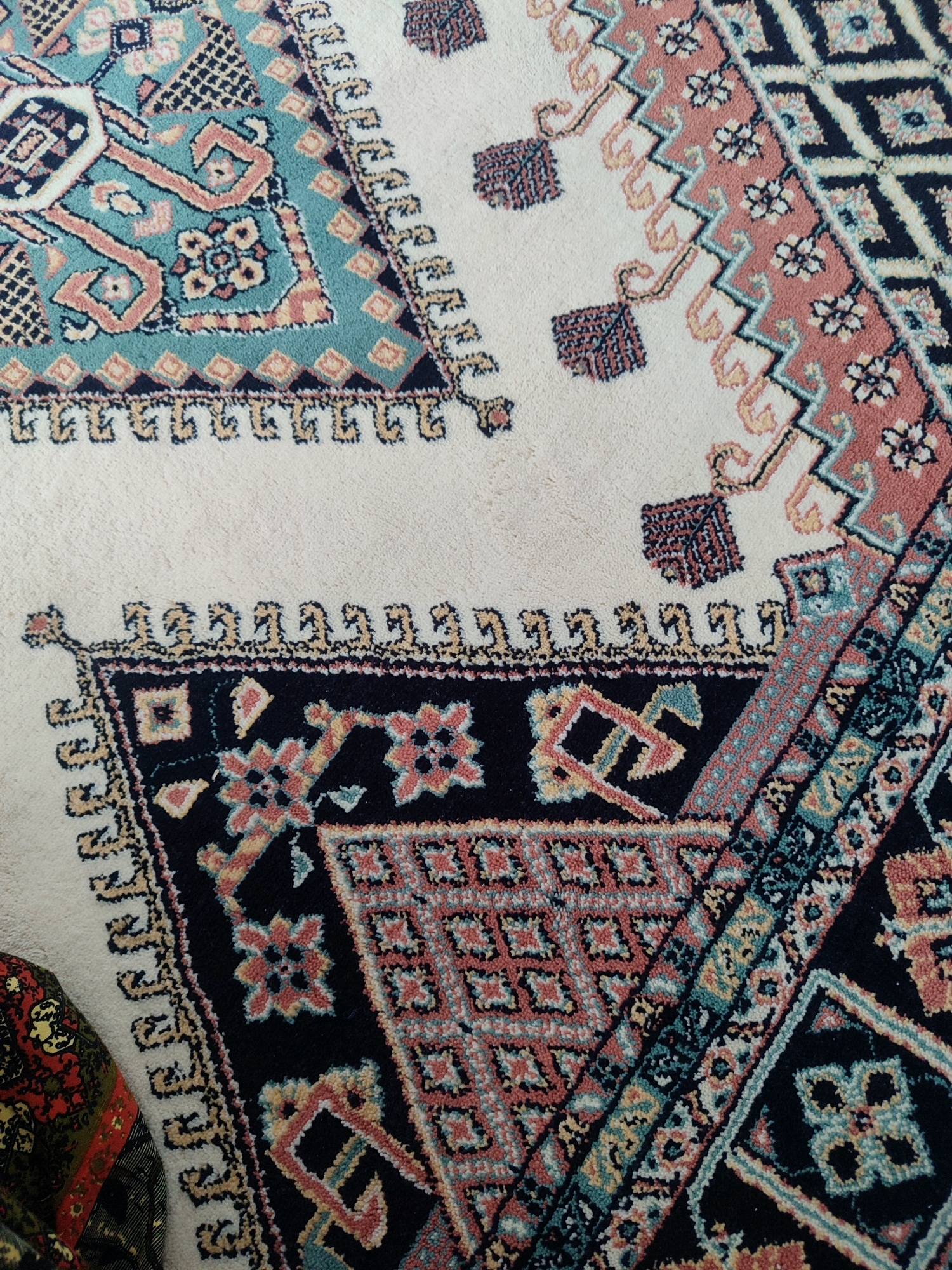 An overhead close-up shot shows a cream-colored rug with a complex geometric pattern featuring teal, coral, navy, and yellow accents. The rug's border is a repeating fringe of small, tan, abstract shapes. In the lower left corner, a portion of a dark, patterned textile is visible, appearing to be another rug or fabric. The image is a still life of textiles.