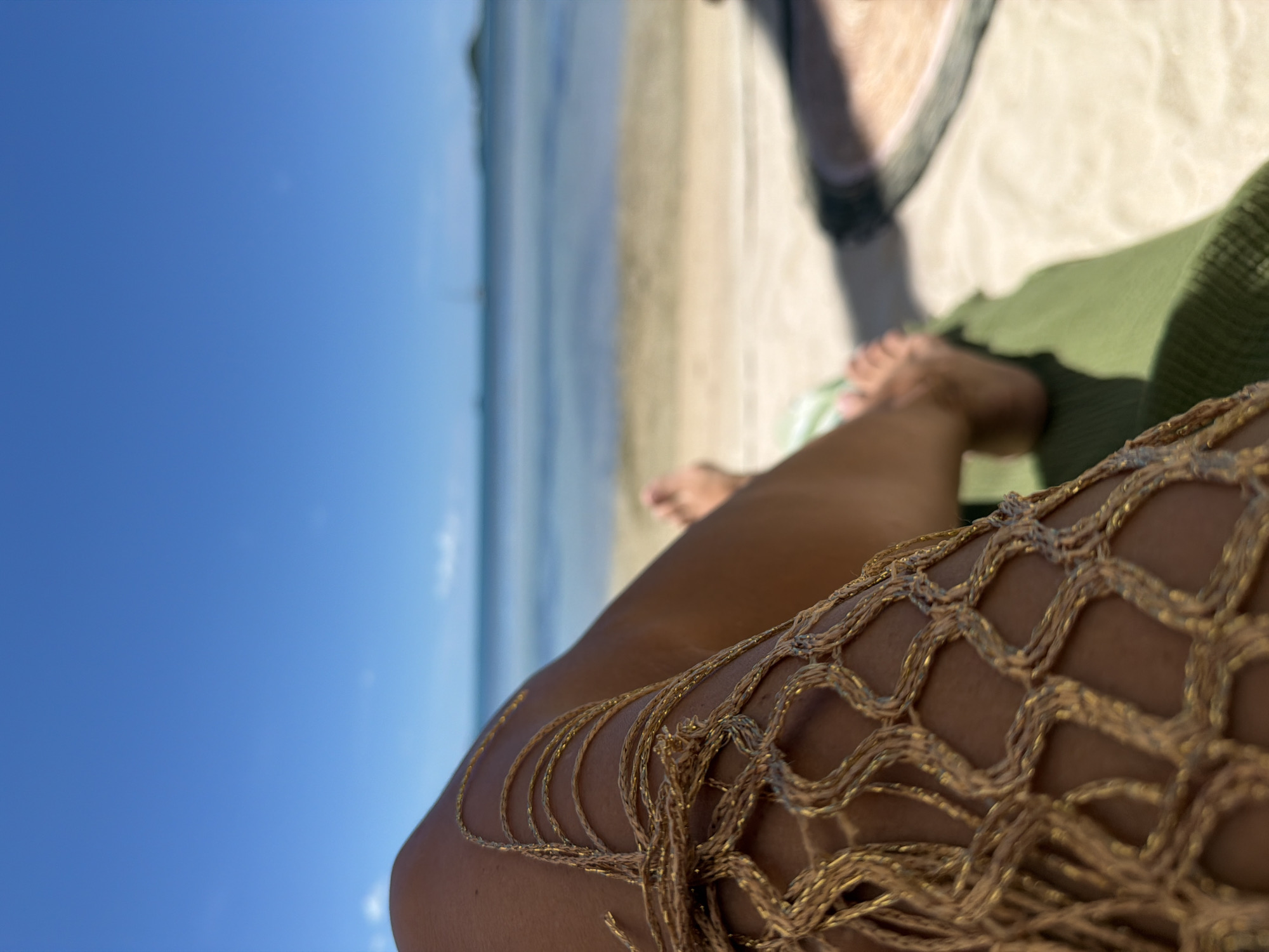 A close-up, over-the-shoulder shot shows a person's bare legs adorned with a gold, netted garment, resting on a green towel. The background is a serene beach scene with the ocean, sand, and a clear blue sky. The location is Baan Thong Krut, Thailand. The water is calm and shallow, with faint outlines of boats visible on the horizon. The sand is light in color. The sun is bright, casting shadows on the sand and the green towel. The focus is sharp on the legs and the netting, while the background is softly blurred, creating a sense of depth and relaxation.