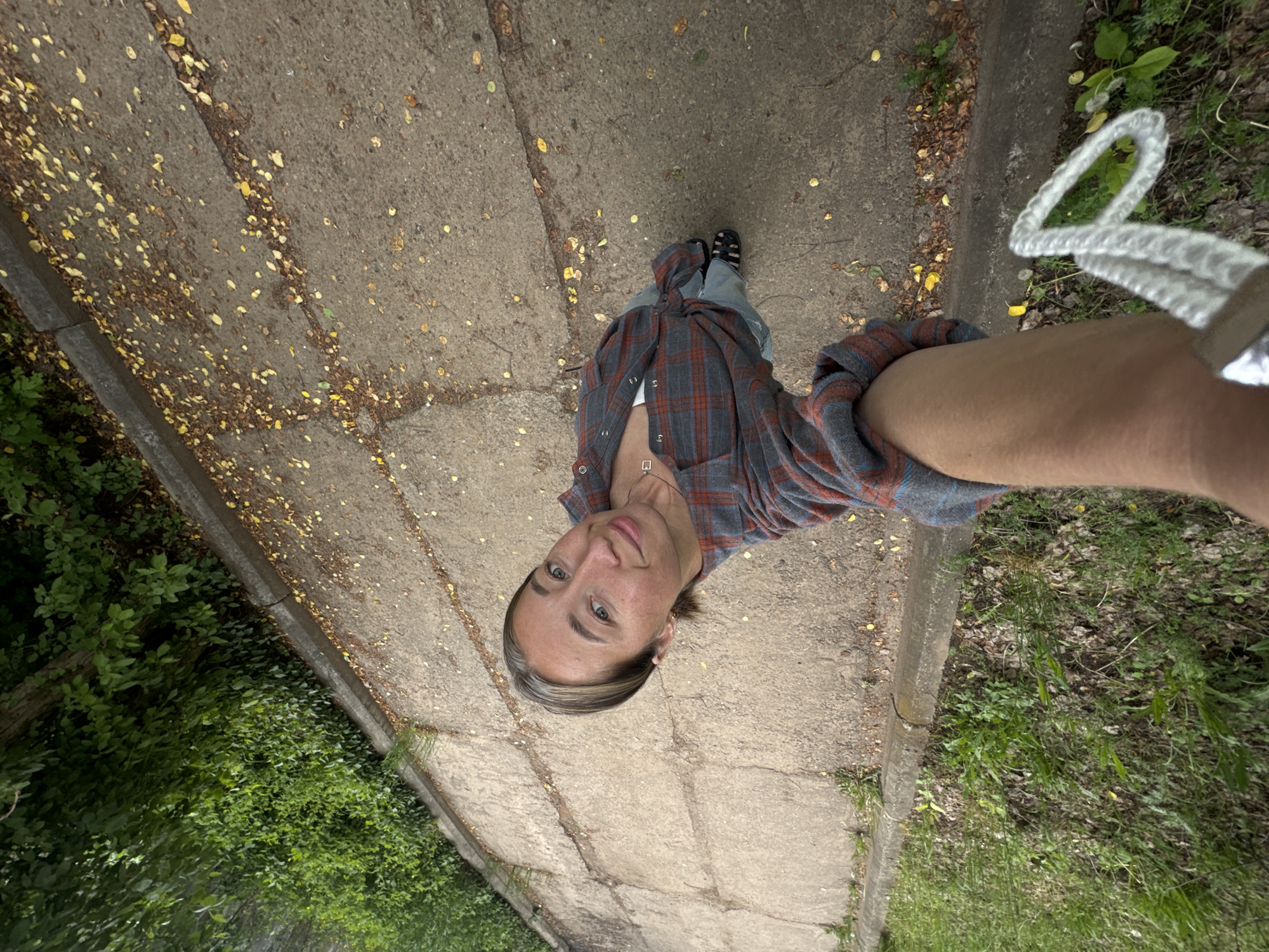 A person is standing on a concrete path, possibly a sidewalk, in an outdoor environment. The camera appears to be held above and at an angle looking down at the person. The person is wearing a plaid shirt over a white top, jeans, and black sandals. The path is cracked and has scattered yellow leaves. To the left of the path is dense green vegetation. To the right, there is more sparse vegetation and what appears to be a white rope or cord held by the person taking the photograph. The angle and lighting suggest a daytime shot. There is no specific evidence in the image to place it in the city of Chaikovsky, Russia.