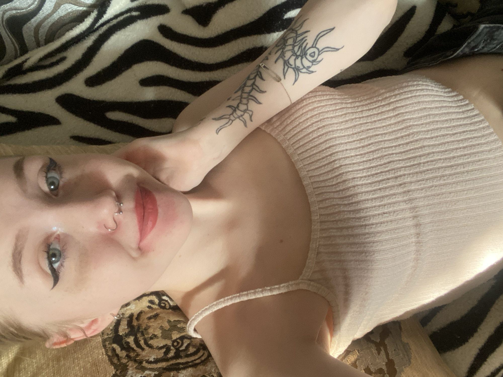 A young woman with light skin, blonde hair, and facial piercings lies on a patterned surface, likely a bed or couch. She wears a light pink tank top and dark-colored bottoms. Her left arm is raised, displaying two insect-like tattoos. The surface beneath her appears to be a zebra-striped fabric and a tiger-striped fabric. There is no indication of Izhevsk, Russia.