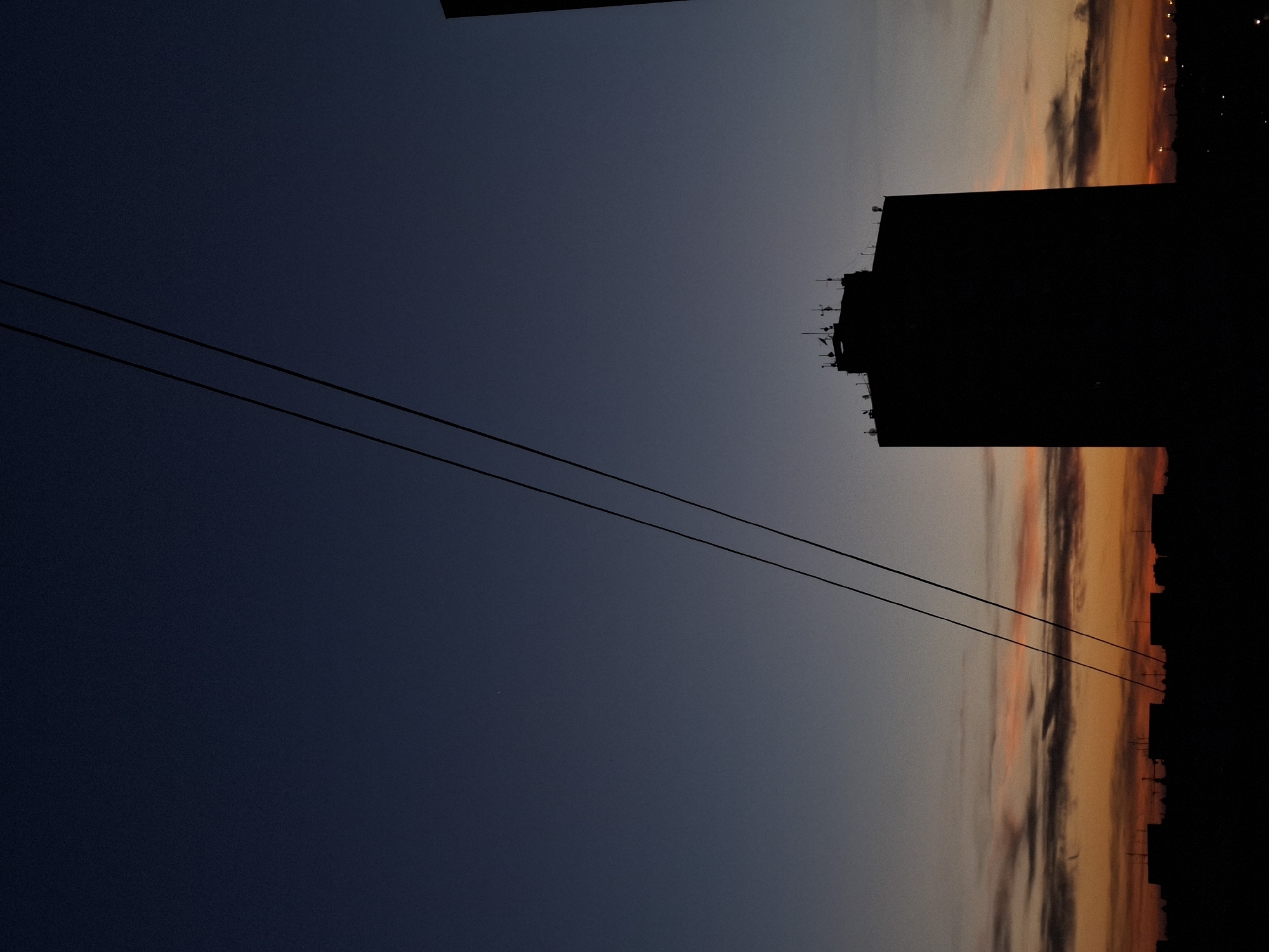 The image is a vertical outdoor shot of a twilight sky in Novi Sad, Serbia. The scene depicts a dark blue sky transitioning to shades of orange and pink near the horizon. Two power lines are visible diagonally across the frame. A silhouetted building with multiple antennas on top is on the right side. The city skyline is also in silhouette along the horizon.