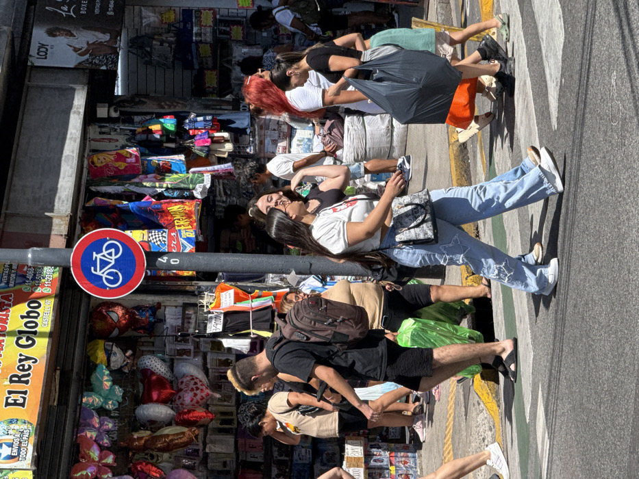 A busy street scene shows people walking alongside shops. A sign with a blue bicycle symbol is visible near a shop named "El Rey Globo". People are browsing the merchandise displayed outside the shops. The image is taken from a street-level perspective, capturing the everyday hustle and bustle of the urban environment.