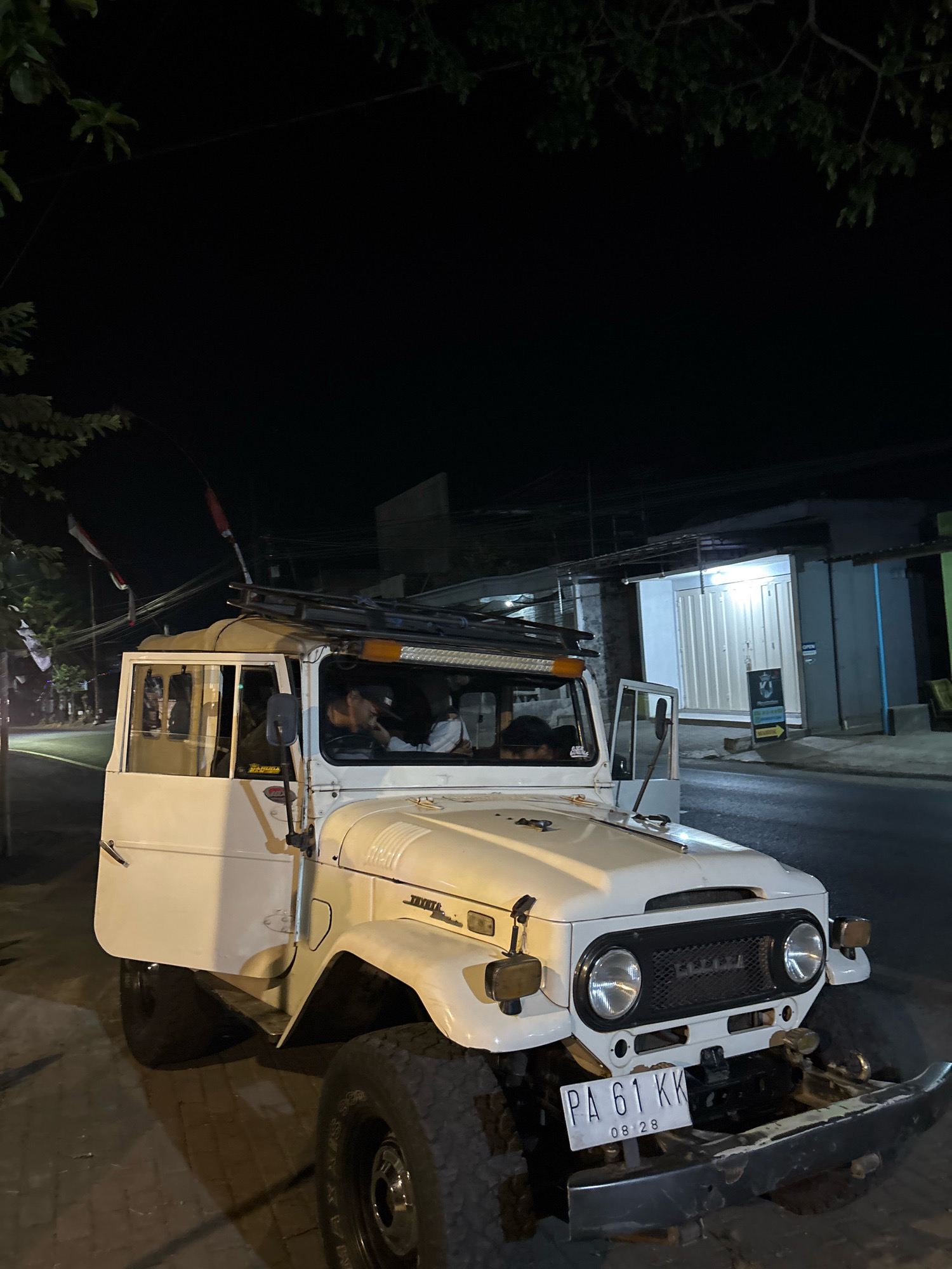 A white Toyota Land Cruiser FJ40 is parked on a brick sidewalk at night. The vehicle has a roof rack and two people are inside. The driver, a male with dark hair, is leaning on his hand. A second person is visible in the passenger seat. Red and white Indonesian flags are attached to poles at the front of the vehicle. In the background, a building with a white, corrugated metal garage door is illuminated. The street is paved, and there are trees and power lines visible. A license plate on the front of the vehicle reads "PA 61 KK" and "08 28". The location is identified as Belung, Indonesia.