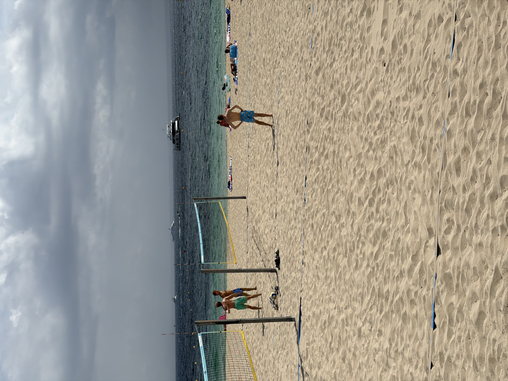 The image depicts a beach scene in Palmanova, Spain, on a cloudy day. In the foreground, sand stretches across the beach, marked by blue lines indicating a volleyball court. Two young men are playing volleyball on the court. In the background, the turquoise sea meets the horizon under a sky filled with dramatic clouds. Several yachts are visible in the distance on the water. Other beachgoers are scattered across the sand, some sunbathing.
