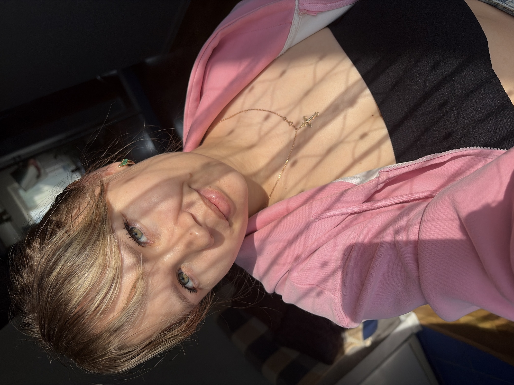 A young woman with blonde hair and green eyes is shown from a low angle, presumably a selfie. She is wearing a pink hoodie over a black top and a delicate gold necklace with a cross pendant. Light and shadow play across her face and body. She appears to be indoors, with dark surfaces visible in the background. Although the location is given as Chaikovsky, Russia, there is no discernible information in the image to verify this.