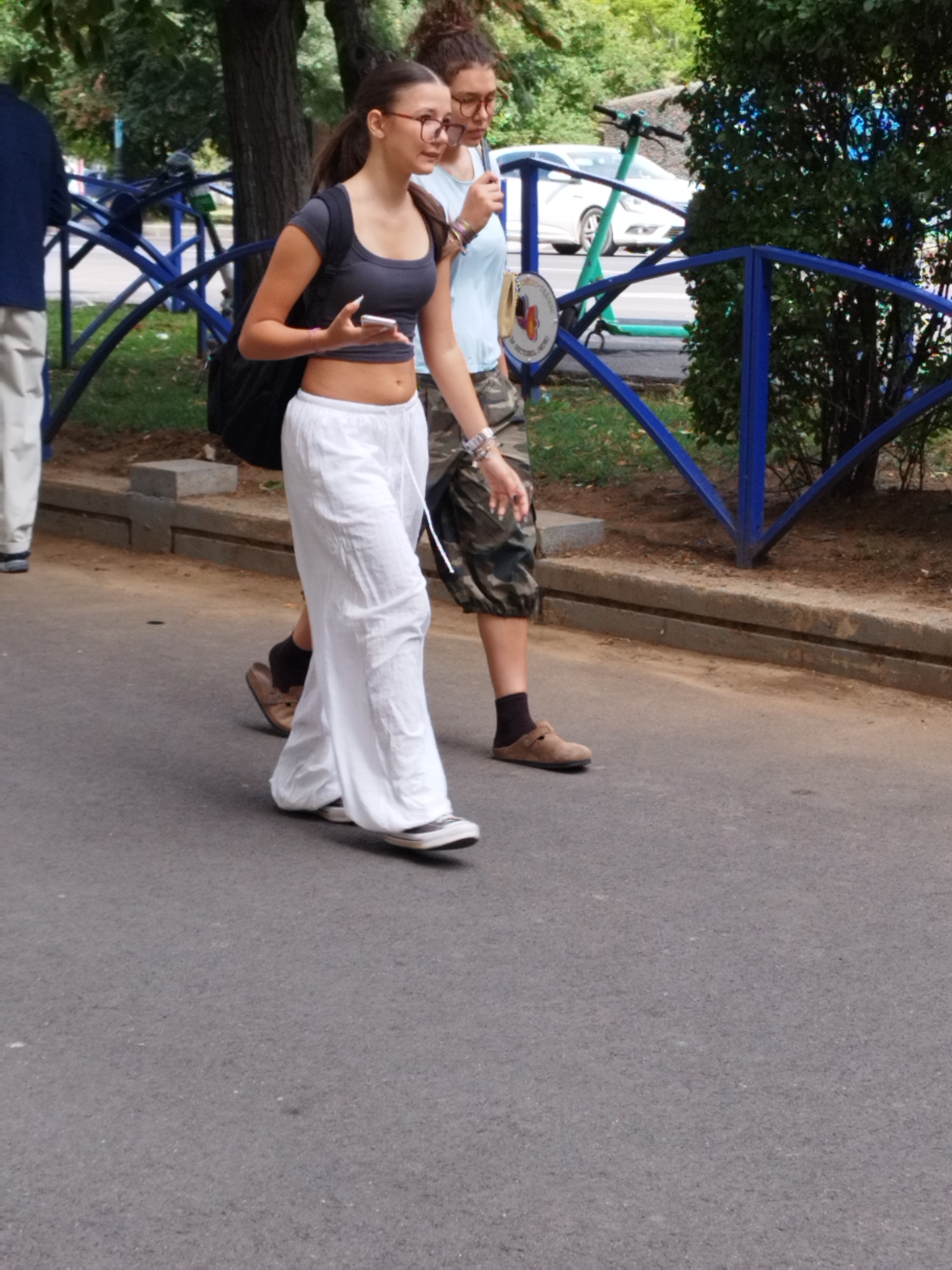 Two young women are walking side-by-side on a paved surface, possibly a sidewalk adjacent to a street. The woman in the foreground, on the left, is wearing a dark grey crop top, a long, flowy white skirt, and black sneakers. She has dark hair pulled back in a ponytail and is wearing glasses and holding a smartphone. The woman behind her, on the right, is wearing a light blue t-shirt and camouflage-patterned shorts. She also wears glasses. Both women are walking towards the left side of the frame. In the background, there are trees, a blue metal fence, and a white car. An electric scooter is visible in the background. The scene appears to be a street in Bucharest, Romania, based on the distinct architecture and vegetation. The lighting suggests it is daytime, with shadows present.