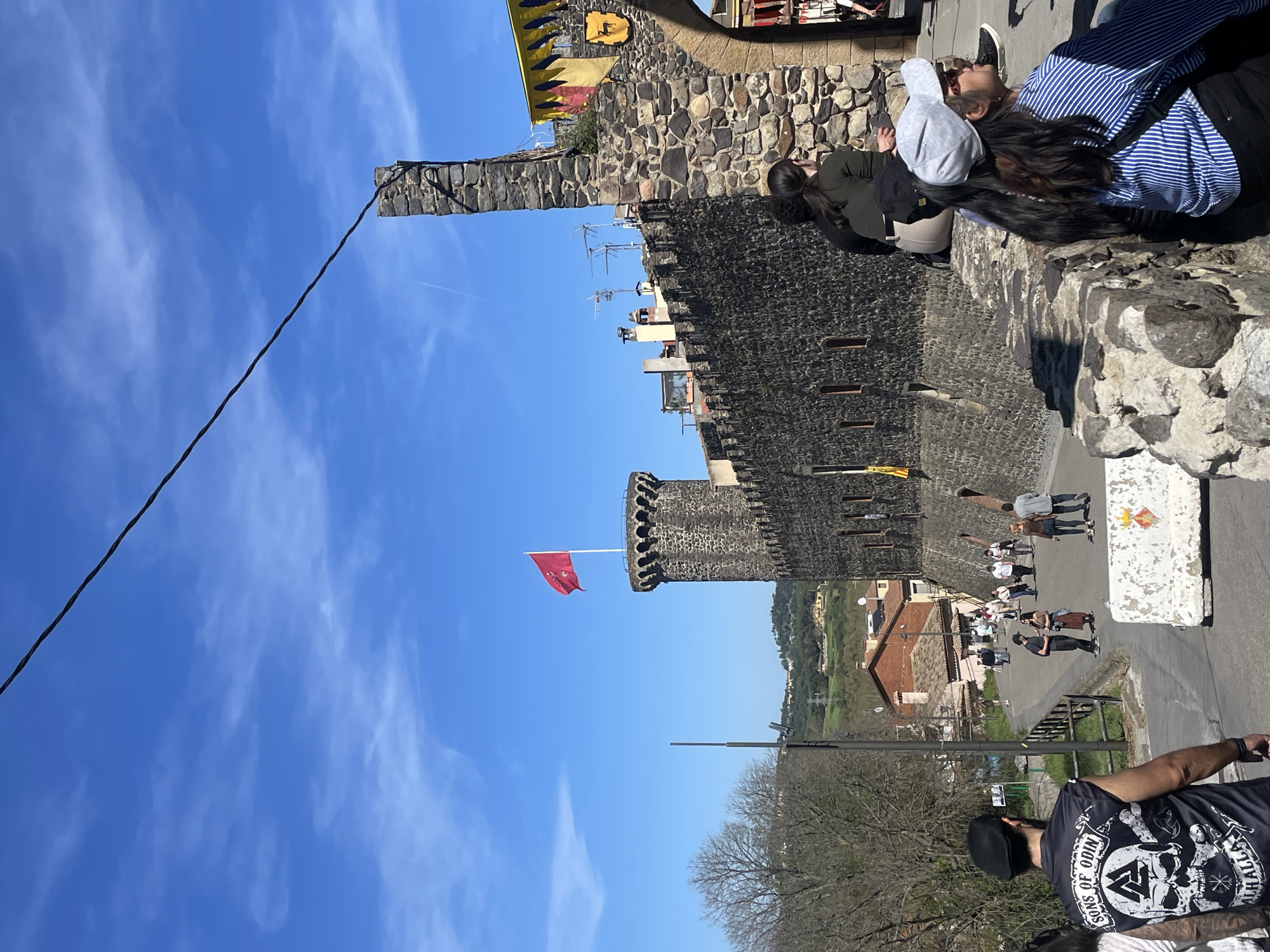 This image depicts Hostalric Castle in Hostalric, Spain. The castle features stone walls, a square tower, and a round tower with a red flag. Tourists are walking along the castle walls, and others are on the path below. The sky is blue with some clouds. A powerline runs through the sky, and a small airplane contrail is visible.