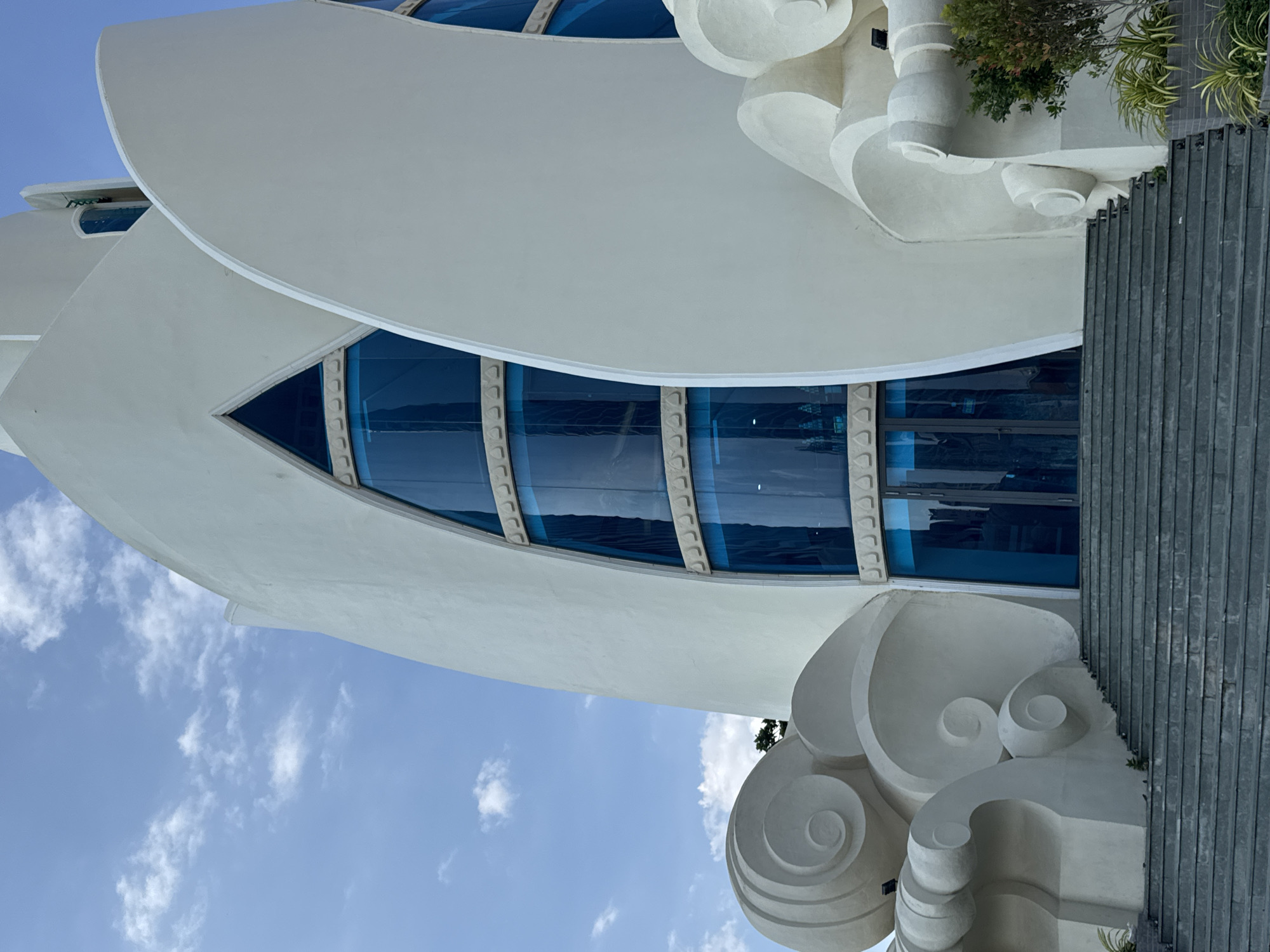 The image depicts the exterior of the Ba Na Hills French Village in Da Nang, Vietnam, specifically showcasing the architecture of one of its prominent buildings. The structure is characterized by its white, organic, and flowing design, resembling stylized waves or petals. Large, arched windows with striking blue glass are a focal point, creating a modern contrast with the sculpted exterior. The building is accessed by a wide, gray stone staircase. The sky above is a bright blue with scattered white clouds, suggesting a daytime scene. Lush green foliage is visible to the right of the staircase, bordering the building. The architectural style appears to be a blend of fantasy and contemporary design, typical of themed attractions.