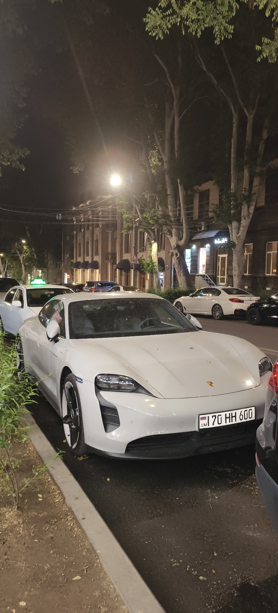 A white 2020 Porsche Taycan 4S is parked on the right side of a street at night in an urban area. The car is facing forward and its front is visible. The license plate reads "170 HH 600". The car is parked next to a curb and a sidewalk with some plants. In the background, there are buildings and trees. Other cars are also visible on the street. A street light is illuminating the scene. The overall impression is a nighttime street scene in a city.