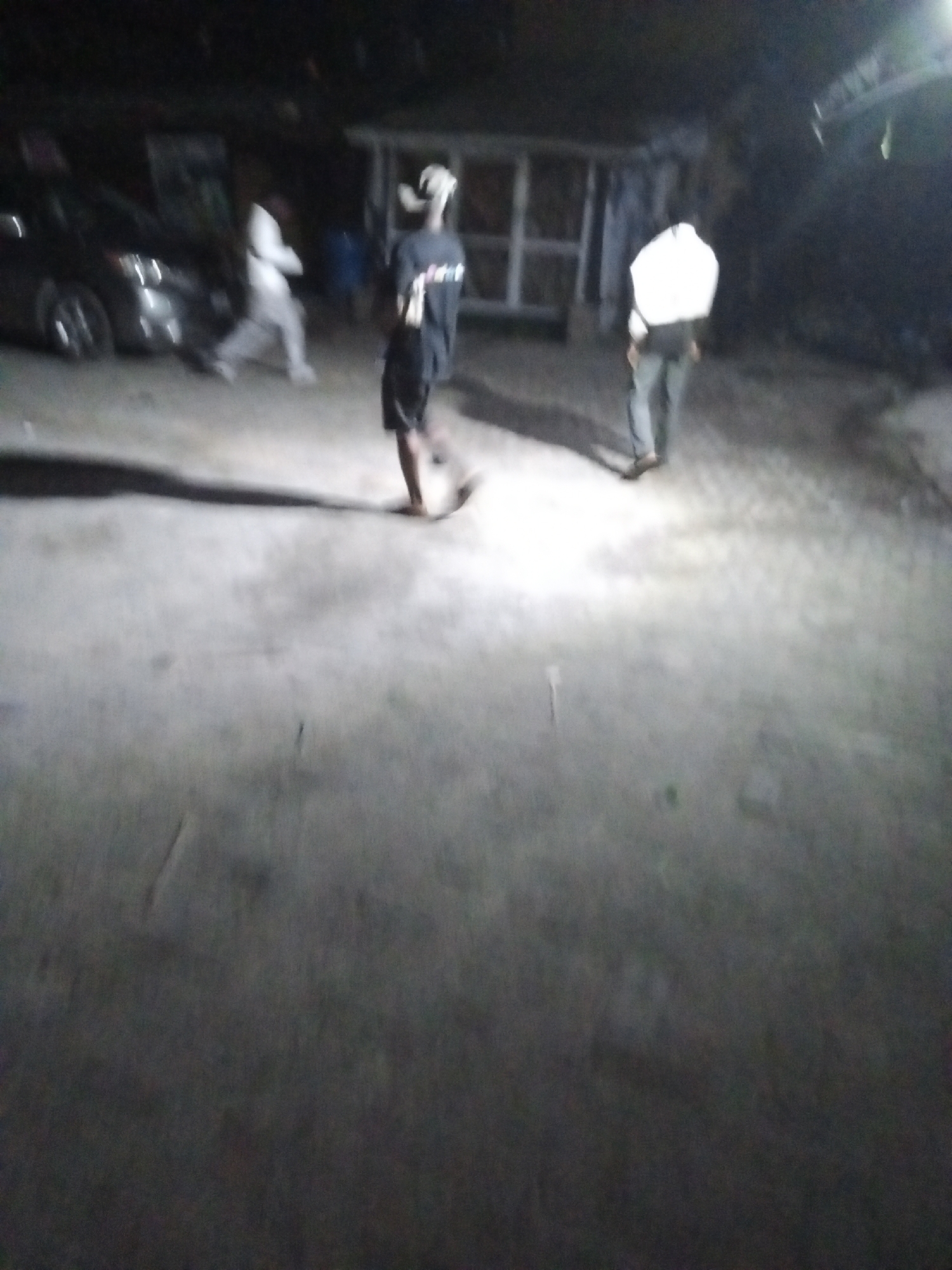 The image was likely taken at night in Ado, Nigeria. The location appears to be an open space or street. There are three people in the frame. One person is wearing a white top and dark trousers and is walking away from the camera. A second person is wearing a dark shirt and shorts and is walking towards the camera. The third person is wearing a white top and light-colored trousers and is walking away from the camera. A car is visible in the background. There is a small, white structure with visible windows or openings also in the background. The ground is a light, solid surface. The scene is generally dark with some areas of brighter illumination.