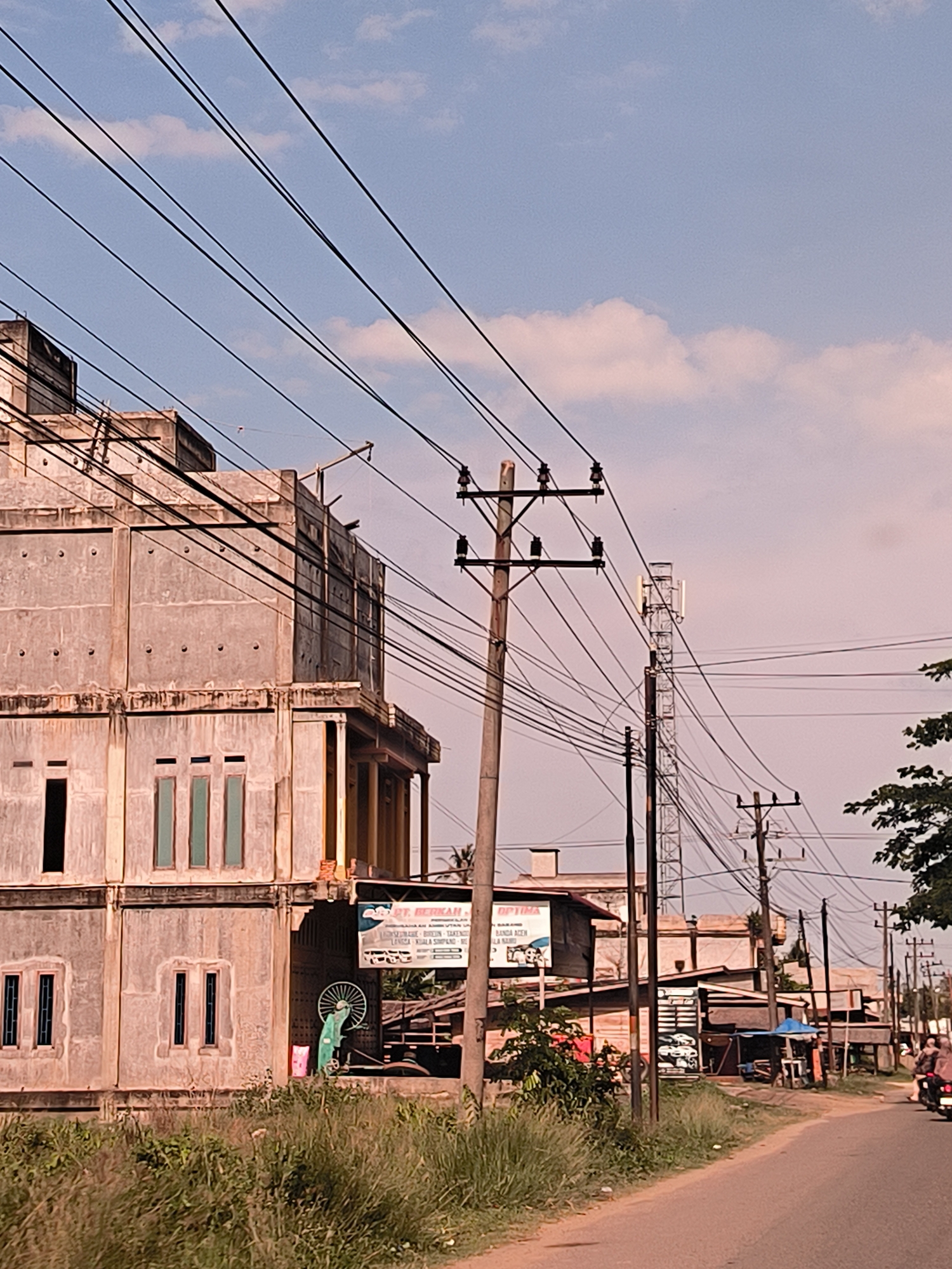 The image depicts a street scene in Teupinbatee, Indonesia. A partially constructed, multi-story concrete building stands prominently on the left. Power lines crisscross overhead, supported by wooden poles. A cell tower is visible further down the street. Vegetation grows along the side of the road. Small commercial establishments line the street, including one with a sign that reads "PT. BERKAH" with further details in Indonesian. A motorcycle with two riders is present on the road. The sky is partially cloudy. The overall environment appears to be a developing or transitional urban area.