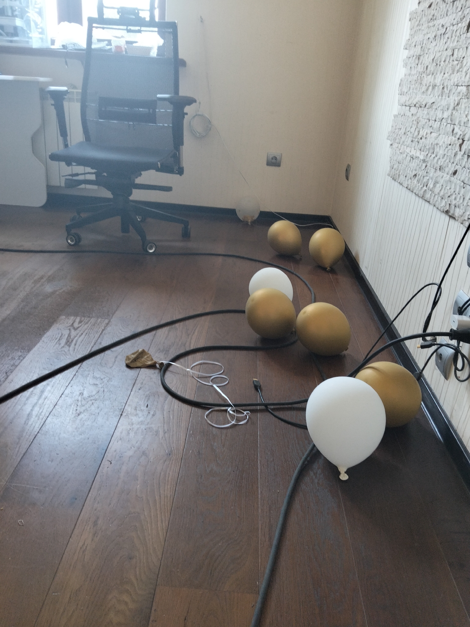An office chair with a black mesh back is positioned on a wooden floor in a room with light-colored walls. Several gold and white balloons are scattered across the floor, some connected by black cords. A tangle of cords and plugs is visible near the wall on the right side of the image. A small piece of crumpled brown paper with a white string attached lies on the floor near a black cord. The scene appears to be indoors, possibly in an office or residential space.