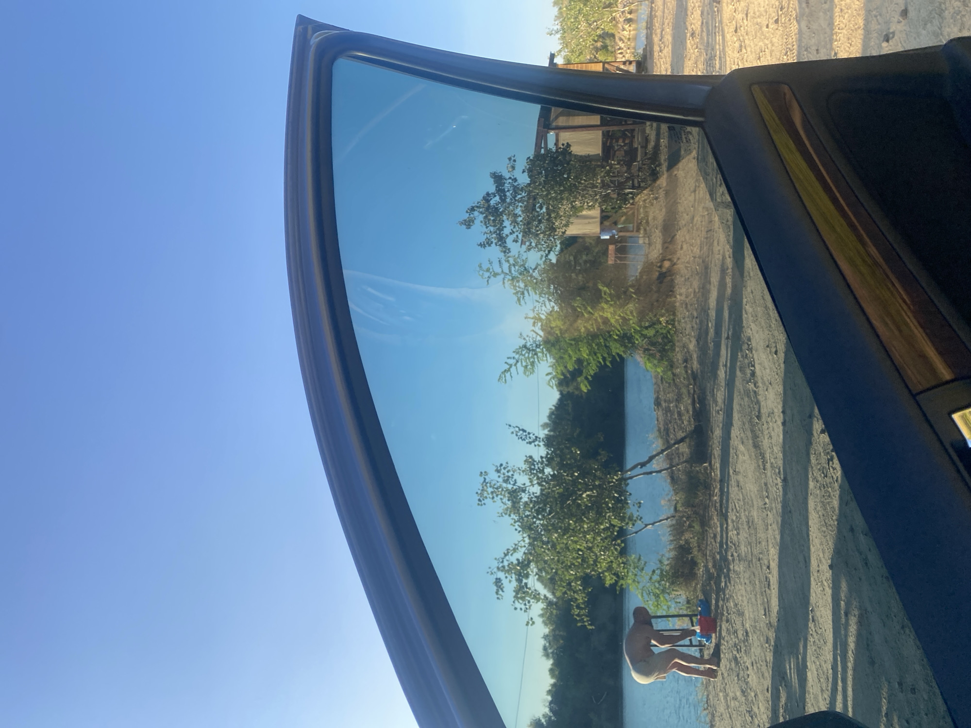 The image depicts a view through a car window, looking out at a body of water with a sandy bank and vegetation. In the foreground, the car's door and tinted window are visible. On the bank, a person, appearing to be male and wearing light-colored shorts, is bent over with their back to the camera. They are near a metal structure and several colorful plastic containers, possibly children's toys. Trees and shrubs are scattered along the bank and in the background, with a small wooden structure visible further down the shore. The sky is clear and blue. Based on the context provided, this scene is located in the city of Dnipro, Ukraine.