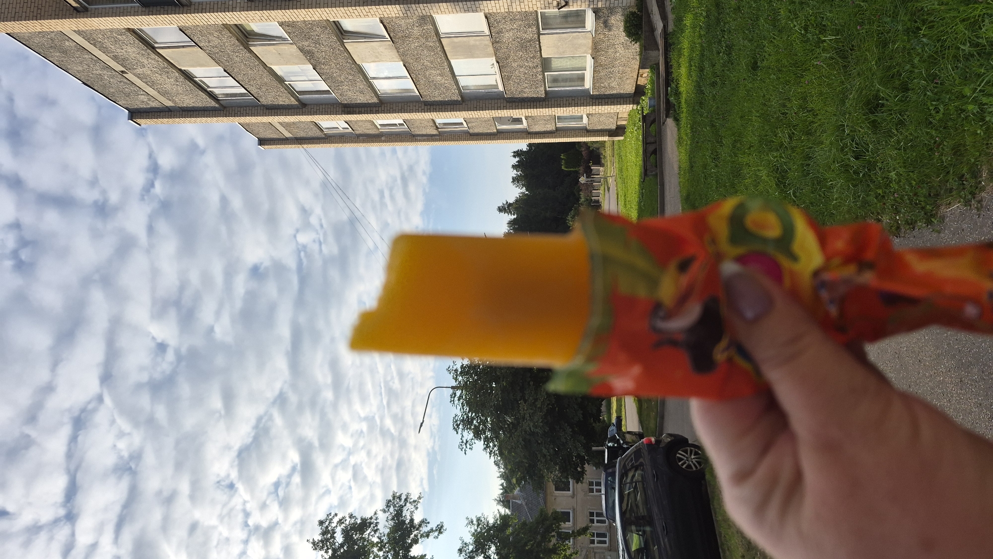 An orange popsicle is held in the foreground by a hand with manicured nails. The popsicle is partially unwrapped, revealing a bite taken from its top. In the background, a multi-story apartment building with beige brick and gray concrete panels dominates the right side of the frame. Adjacent to the building is a well-maintained green lawn with a few wooden benches. To the left of the building, trees and houses are visible, suggesting a residential area. A street and a parked black car are also present on the left side. The sky is filled with scattered white clouds against a blue backdrop. The image appears to have been taken outdoors in a city, possibly in Krauja, Latvia, given the architectural style.