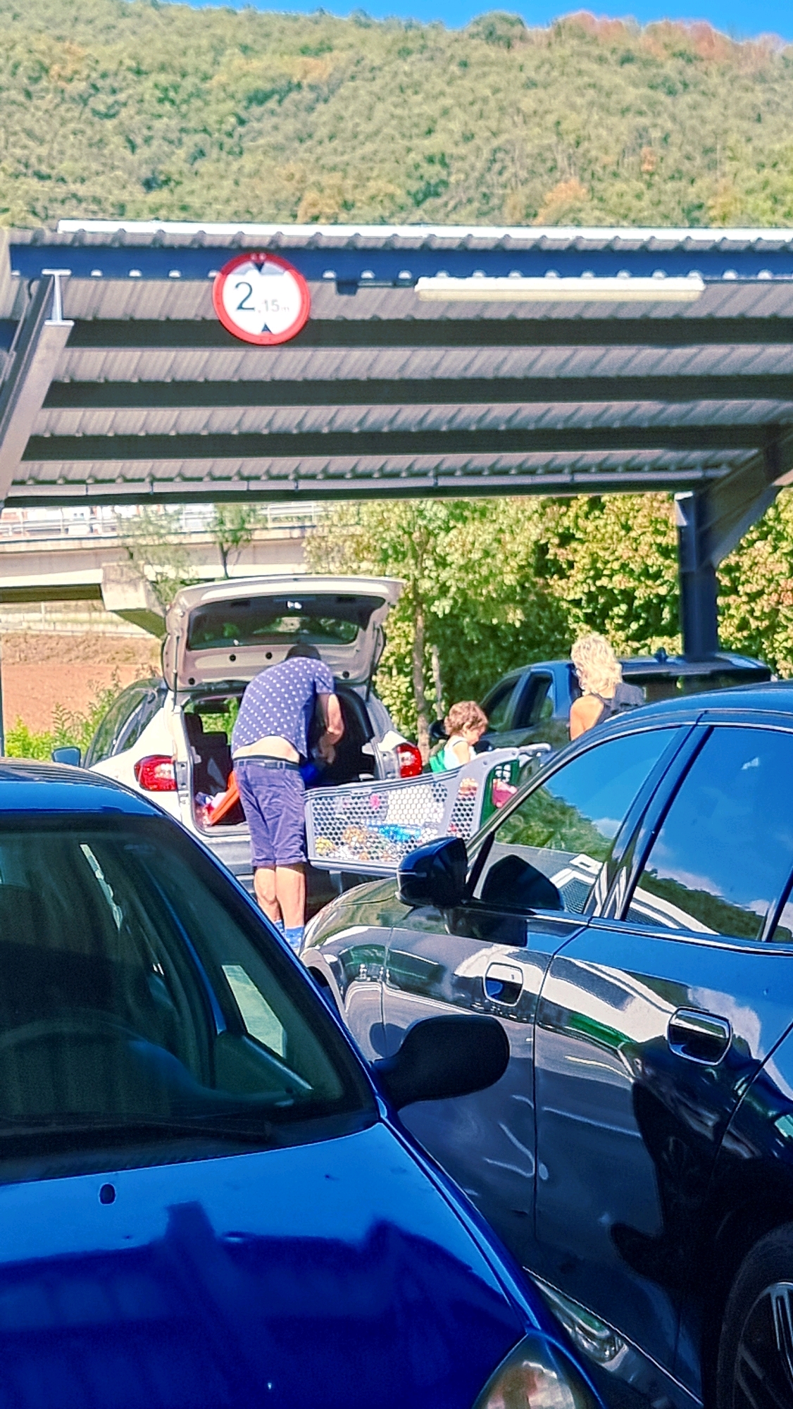 The image depicts a scene under a covered parking structure, likely at a supermarket in Olot, Spain, given the presence of shopping carts and people unloading groceries. In the foreground, the hood of a blue car is visible. Behind it, a dark-colored car is parked, with its side mirror also in view. Further back, a white hatchback with its trunk open is the central focus. A man wearing a blue and white polka-dot shirt and blue shorts is bending into the trunk, attending to a shopping cart filled with groceries. To the right of the white car, a woman with blonde hair is standing, and a young child is visible behind the shopping cart. A road and a hillside with green foliage are visible in the background. A circular red and white sign with "2.15m" is affixed to the roof structure, indicating a height restriction. The overall atmosphere suggests a typical shopping trip with a family unloading their purchases.