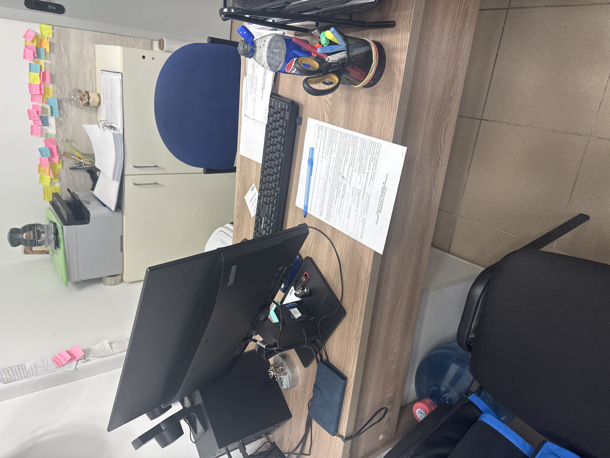 The image shows an office desk in what appears to be Veliko Tarnovo, Bulgaria based on the text in the documents. The desk is cluttered with typical office supplies, including a keyboard, monitor, pens, scissors, and a water bottle. There's a document with Bulgarian text, possibly a protocol for advertising. A notebook and a blue pouch lay near the monitor base. A blue office chair and a blue water bottle are partially visible on the right. On the left, there are cabinets and a wall covered with sticky notes.