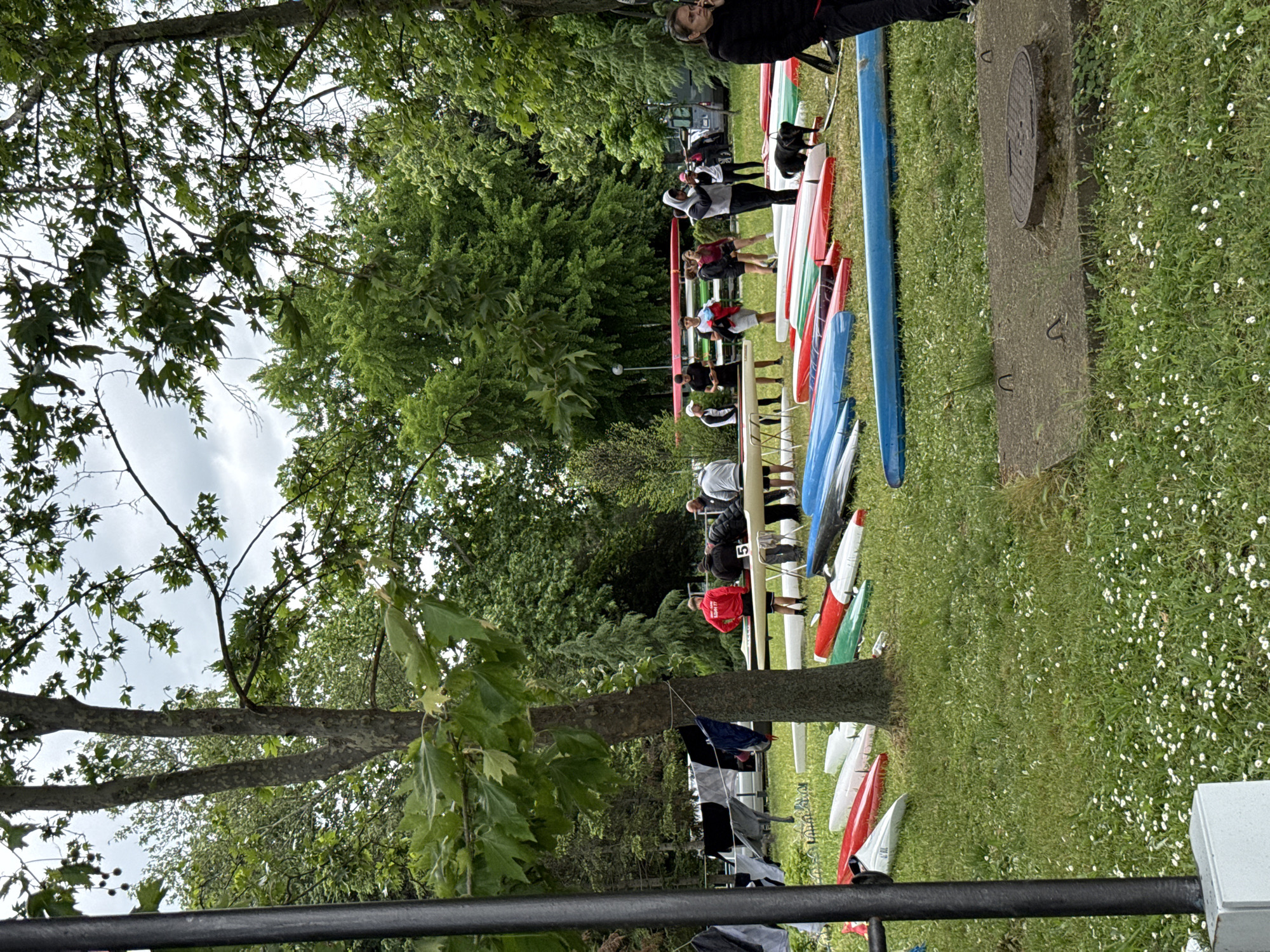 The image depicts a riverside area, potentially in Plovdiv, Bulgaria, given the context, although specific city landmarks are not visible. A group of people, mainly young adults, are gathered around numerous kayaks and canoes laid out on a grassy bank beside a body of water. The boats are of various colors, including red, white, blue, and green. Some individuals appear to be preparing for or finishing a water activity, with some wearing athletic attire. A man with glasses is visible. Trees border the area, suggesting a park or recreational space. A metal railing is in the foreground.
