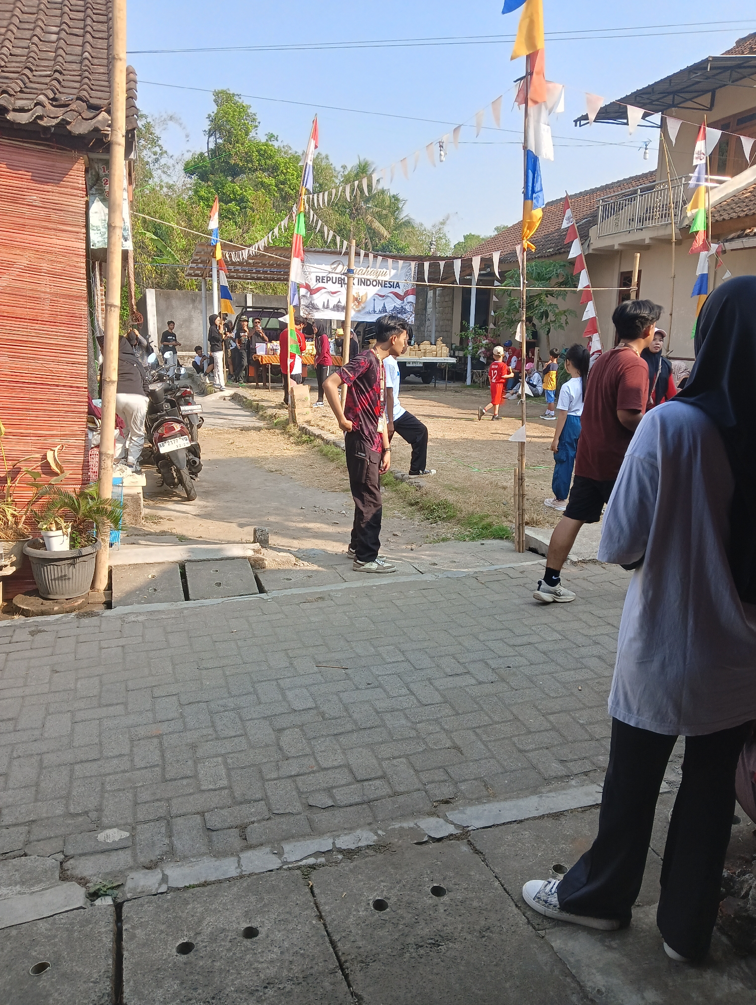 The image captures an outdoor street scene in Mendut, Indonesia, likely during a community event or celebration. The foreground is dominated by a gray brick-paved walkway, with concrete drainage grates visible. Several people are present, some standing and observing, while others appear to be participating in activities or socializing.
In the mid-ground, a pathway leads towards a gathering of stalls or booths, suggesting a market or festival. Various colorful flags, including the Indonesian flag, are displayed, along with decorative pennant banners. A motorcycle is parked to the left, near a red-walled building with bamboo accents.
Further back, more buildings are visible, with rooftops characteristic of Indonesian architecture. Trees and lush greenery are interspersed throughout the background, indicating a tropical environment. The sky is clear and blue, suggesting daylight with ample sunshine.
A variety of individuals are depicted, dressed in casual attire. Some are wearing t-shirts, while others have on more traditional clothing or head coverings. Their actions are varied: some are looking towards the event, one person is standing with hands on hips, and another appears to be holding something in their hand. Interactions seem relaxed and informal. The overall impression is one of a lively, community-oriented gathering in a semi-urban, tropical setting.