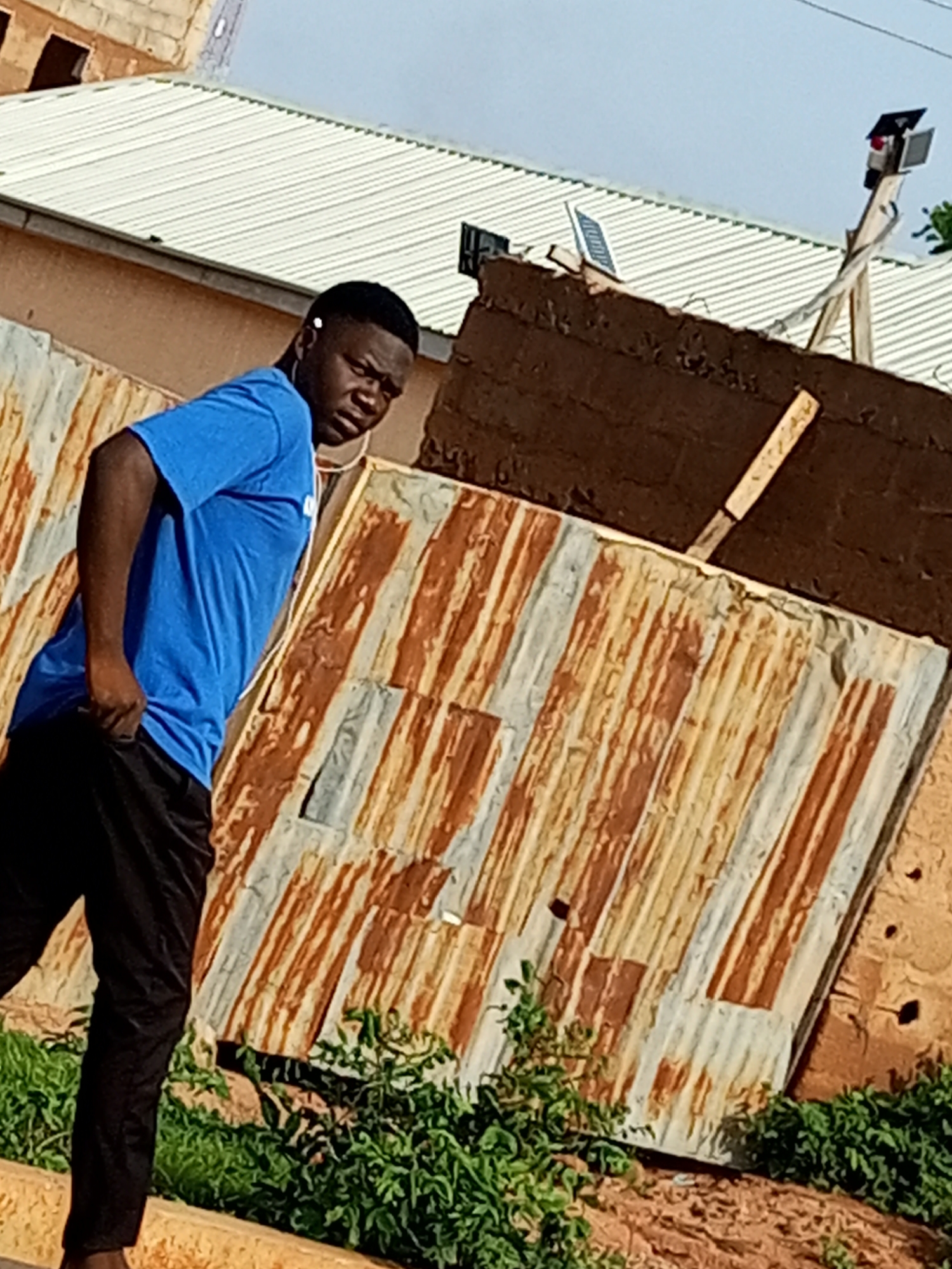 A young man wearing a blue t-shirt and black pants stands outdoors, looking to his right. He has an earbud in his left ear and is holding the front of his pants with his left hand. Behind him, a corrugated metal fence shows signs of rust and decay. In the background, a building with a white, corrugated metal roof is visible, along with a solar-powered light post. The scene appears to be in a rural or semi-urban area, possibly Tumfure, Nigeria, given the environmental context. The ground is covered with vegetation and dirt. The lighting suggests it is daytime, with clear skies.