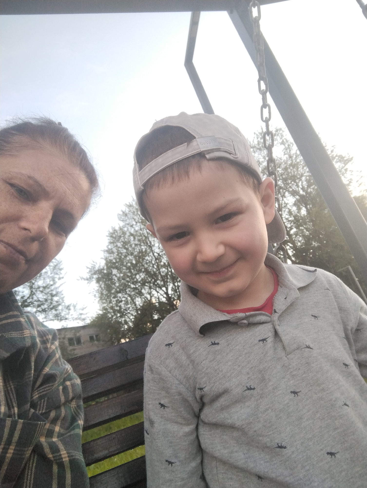 In this close-up, waist-up selfie, a woman and a young boy are positioned in the foreground, appearing to be on a swing set. The woman is on the left, her face turned towards the camera with a neutral expression, her hair pulled back. She is wearing a dark green and tan plaid shirt. To her right, the boy is also facing the camera, smiling broadly. He is wearing a light gray polo shirt with a subtle pattern of small, dark animal figures and a beige baseball cap. Behind them, a wooden bench is visible, and beyond that, trees and a building with windows can be seen. The background suggests an outdoor setting, possibly a park or a yard, with the sky overhead. The image quality is slightly grainy, with a bright sky dominating the upper portion. The location is identified as Kingisepp, Russia.