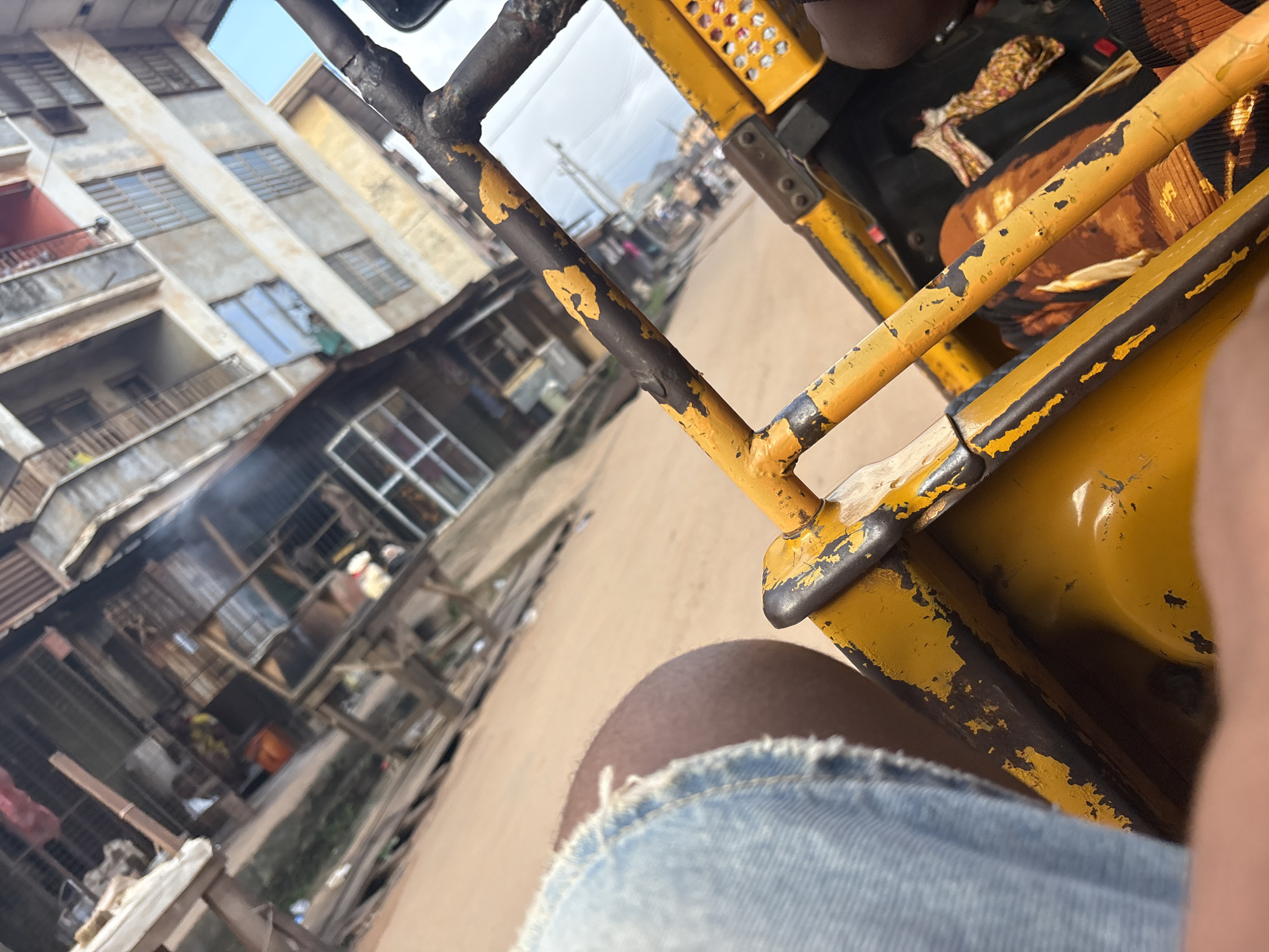 A first-person perspective shot from inside a yellow auto-rickshaw, likely in Onitsha, Nigeria, captures a street scene. The rickshaw's weathered, peeling yellow and black paint is prominent in the foreground. To the left, a person's bare leg wearing distressed denim shorts is visible. In the background, a narrow street is lined with buildings of varying heights and states of repair, some appearing to be shops or residences with visible windows and corrugated metal roofs. Smoke or steam rises from one of the structures, suggesting activity like cooking or manufacturing. The ground appears to be a dusty, sandy road. The overall atmosphere suggests a bustling, urban environment.