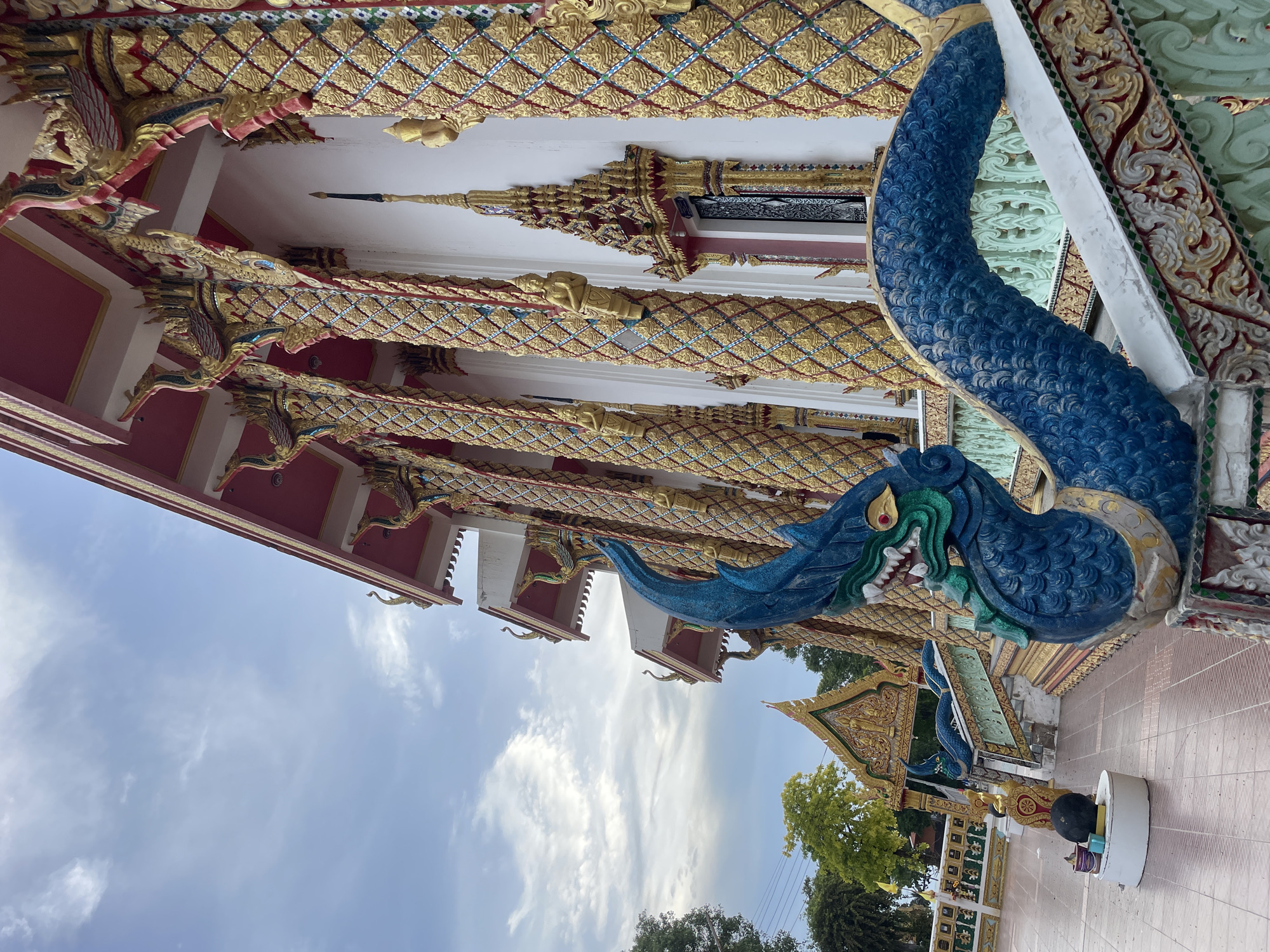 The image shows the exterior of a Buddhist temple in Pattaya City, Thailand. A prominent blue, serpentine dragon sculpture, likely a Naga, is positioned on the left side of the frame, forming part of a staircase railing. The temple building itself is ornate, with a white facade adorned with numerous elaborately decorated golden pillars featuring intricate patterns and small golden figures. The roofline is red, with decorative elements also in gold and red. The sky above is partly cloudy, with a mix of blue sky and white clouds. In the background, there are trees and other temple structures, including an arched gateway with a golden roof. The ground appears to be paved with light brown tiles. There are no people visible in the image.